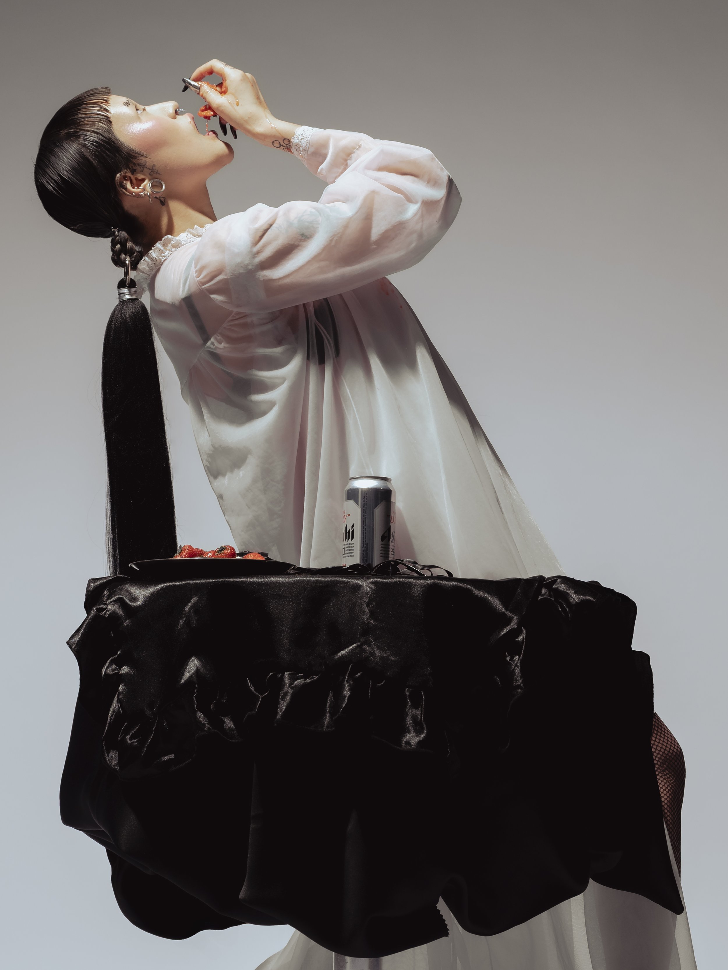 Une femme avec une coiffure longue, portant un vêtement blanc, mangeant des fraises et une canette de boisson est debout derrière une table noire en satin.