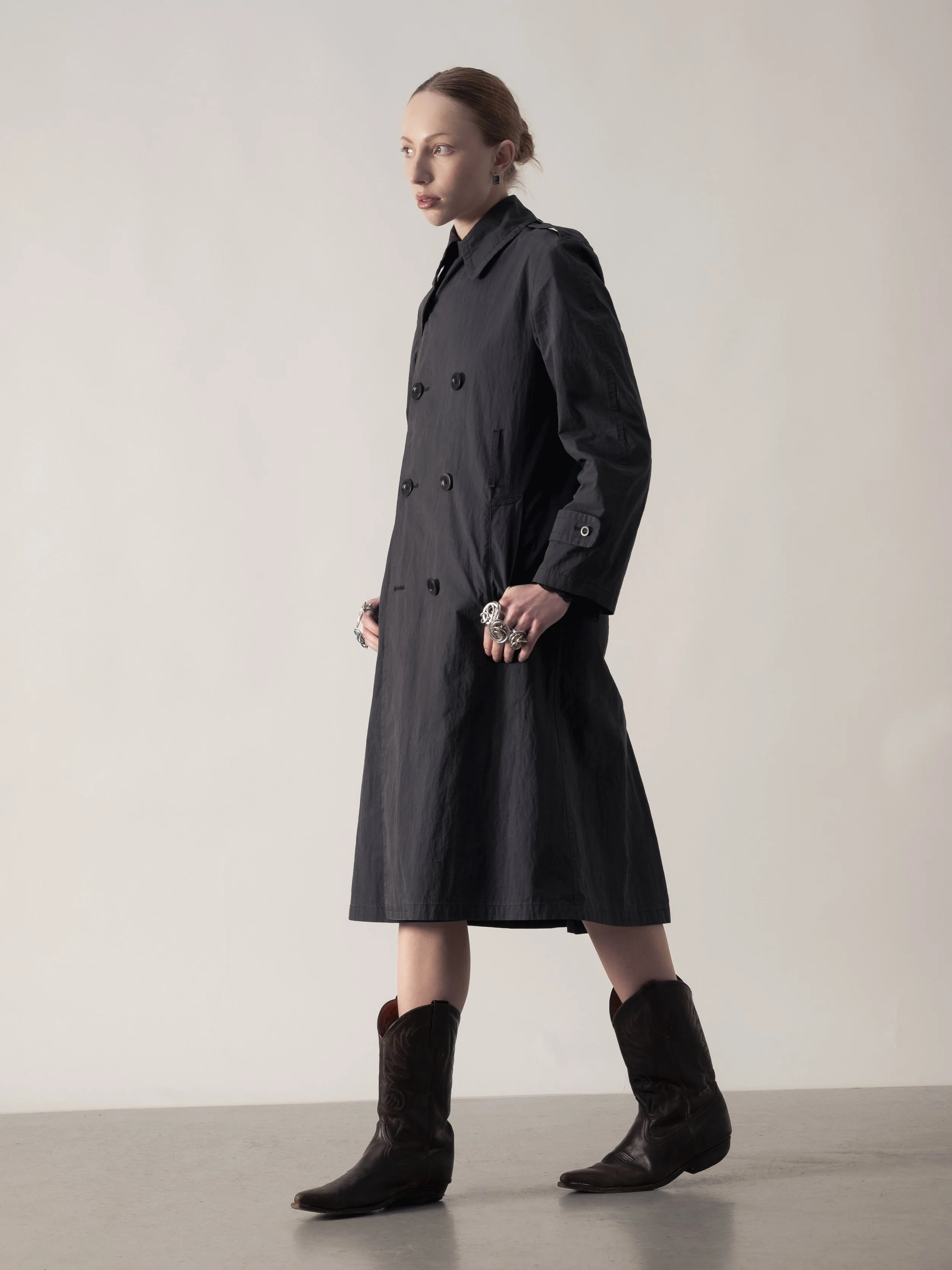 Une femme portant un trench-coat noir et des bottes en cuir marron clair, debout sur un sol gris, vue de profil, contre un fond neutre.