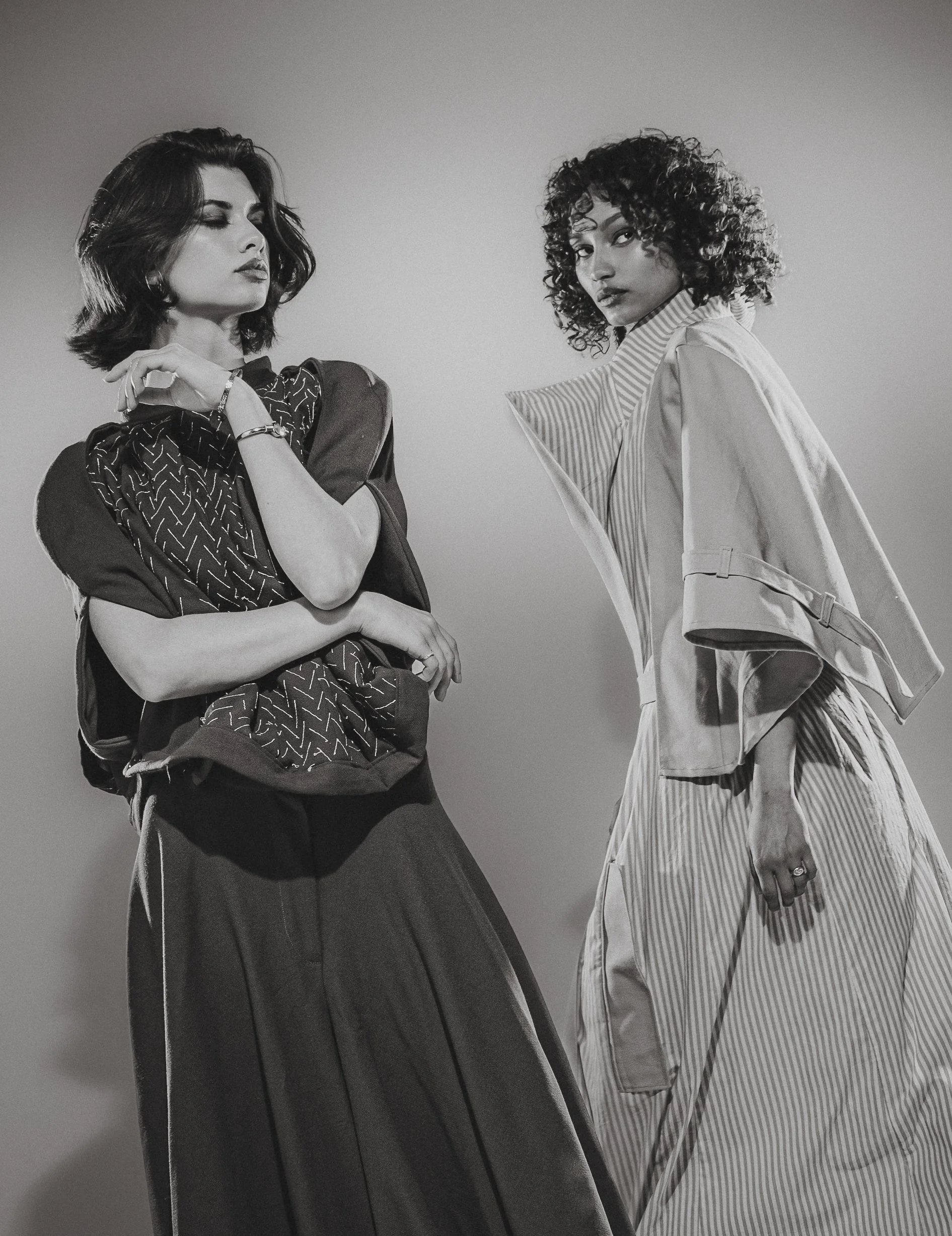 Deux femmes posent pour une séance photo, l'une avec des cheveux courts et foncés, l'autre avec des cheveux bouclés, portant des vêtements à motifs et des vêtements de style décontracté. La photo est en noir et blanc.