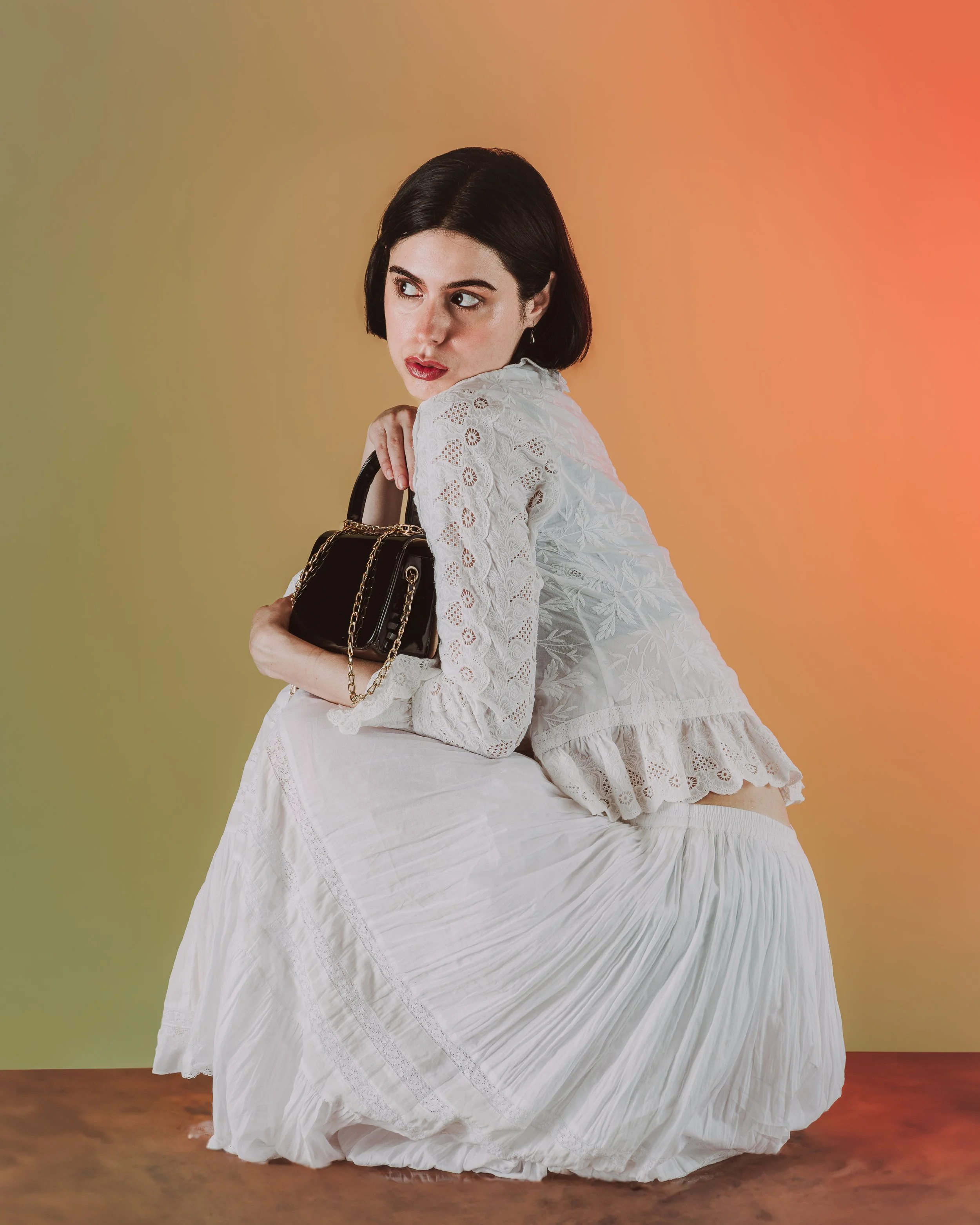 Jeune femme avec une robe en dentelle blanche et une jupe plissée blanche, tenant un petit sac à main noir avec chaîne dorée, posant contre un fond dégradé de couleurs pastel.