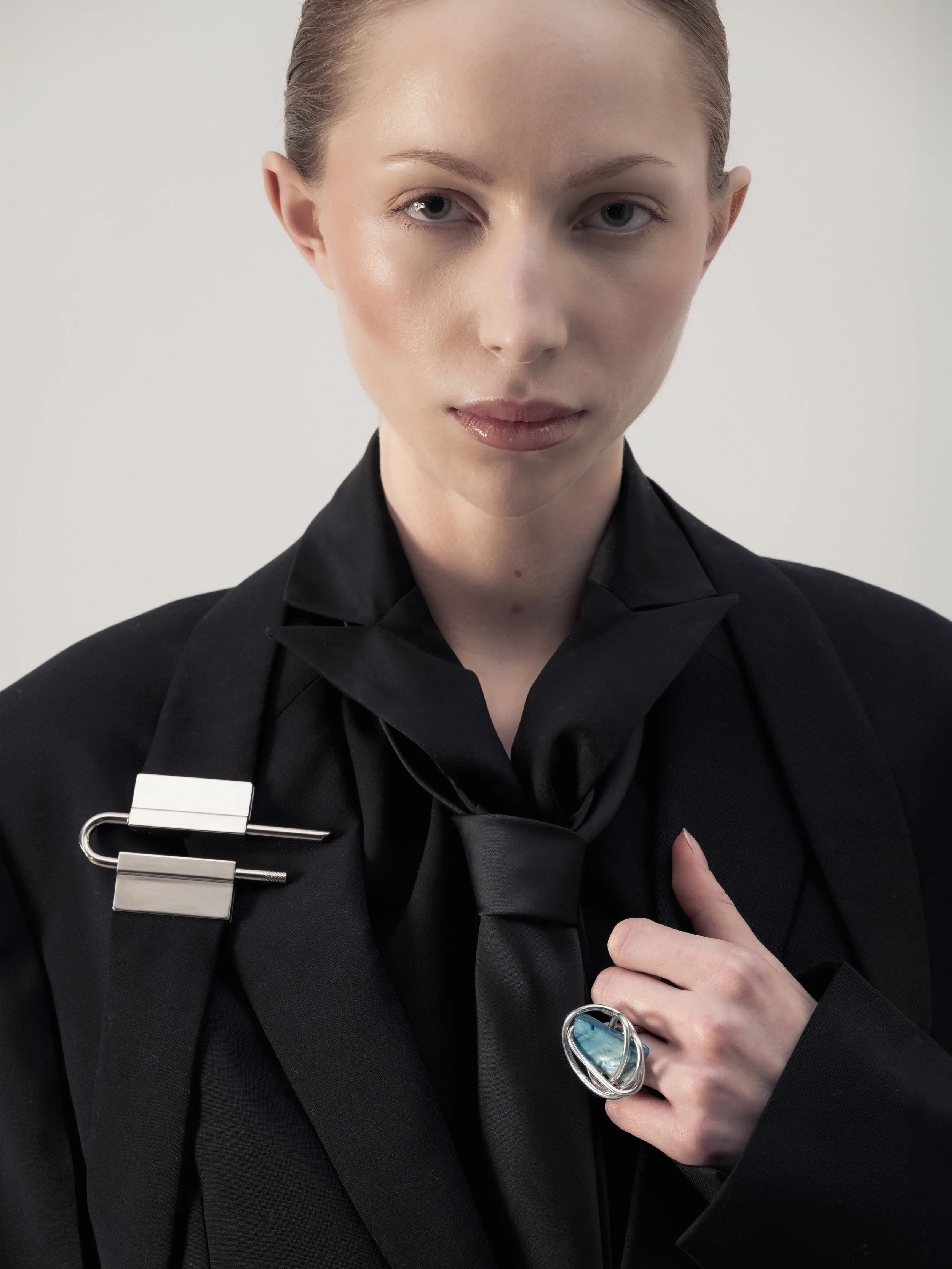 Femme habillée en noir avec un look sophistiqué, portant une grande bague bleue et un broche argentée sur une épaule.
