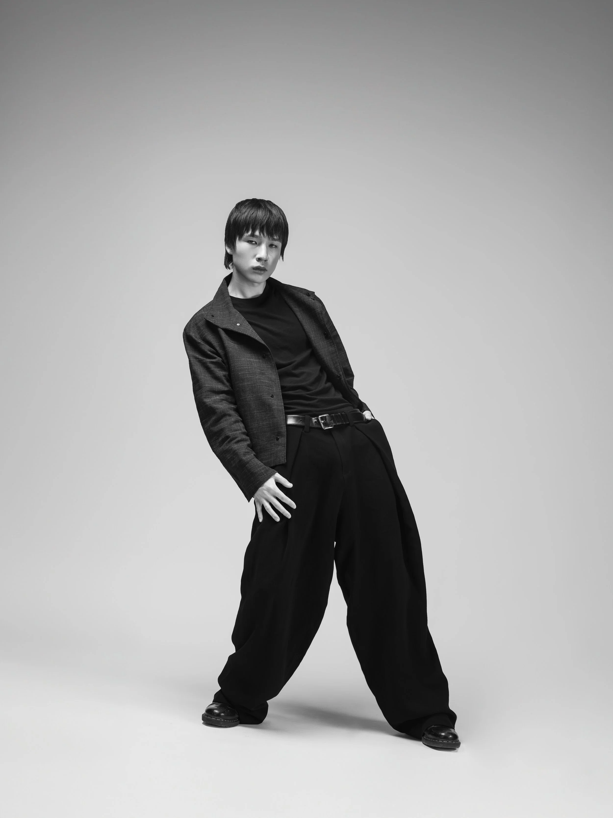 Une personne posant dans une tenue décontractée noire et une veste en denim, sur un fond gris. La personne a les cheveux courts et une expression neutre.