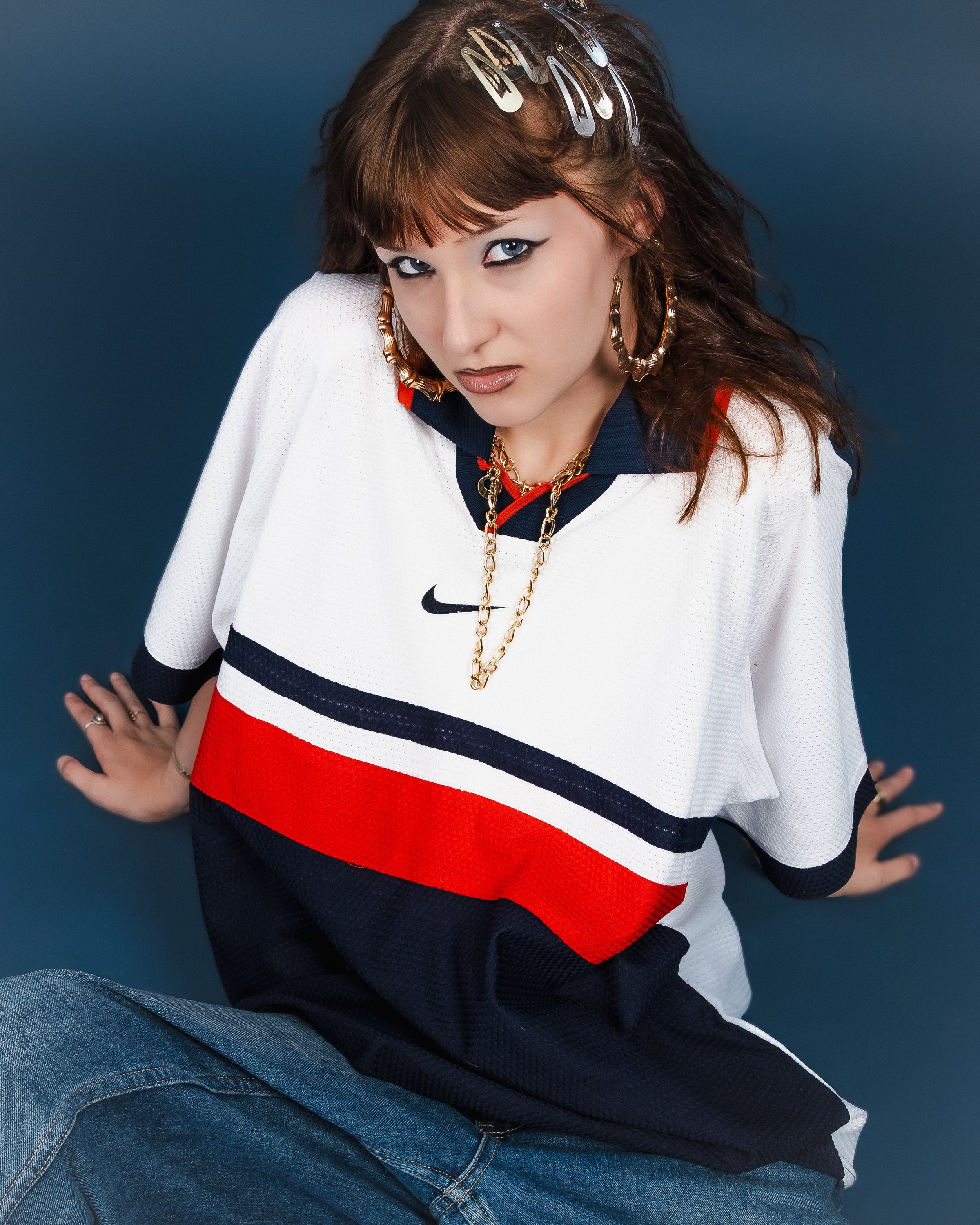 Une femme portant un maillot de sport Nike blanc avec des bandes rouge et noire, des boucles d'oreilles en or, un collier en or, et des pinces à cheveux argentées, posant contre un fond bleu.