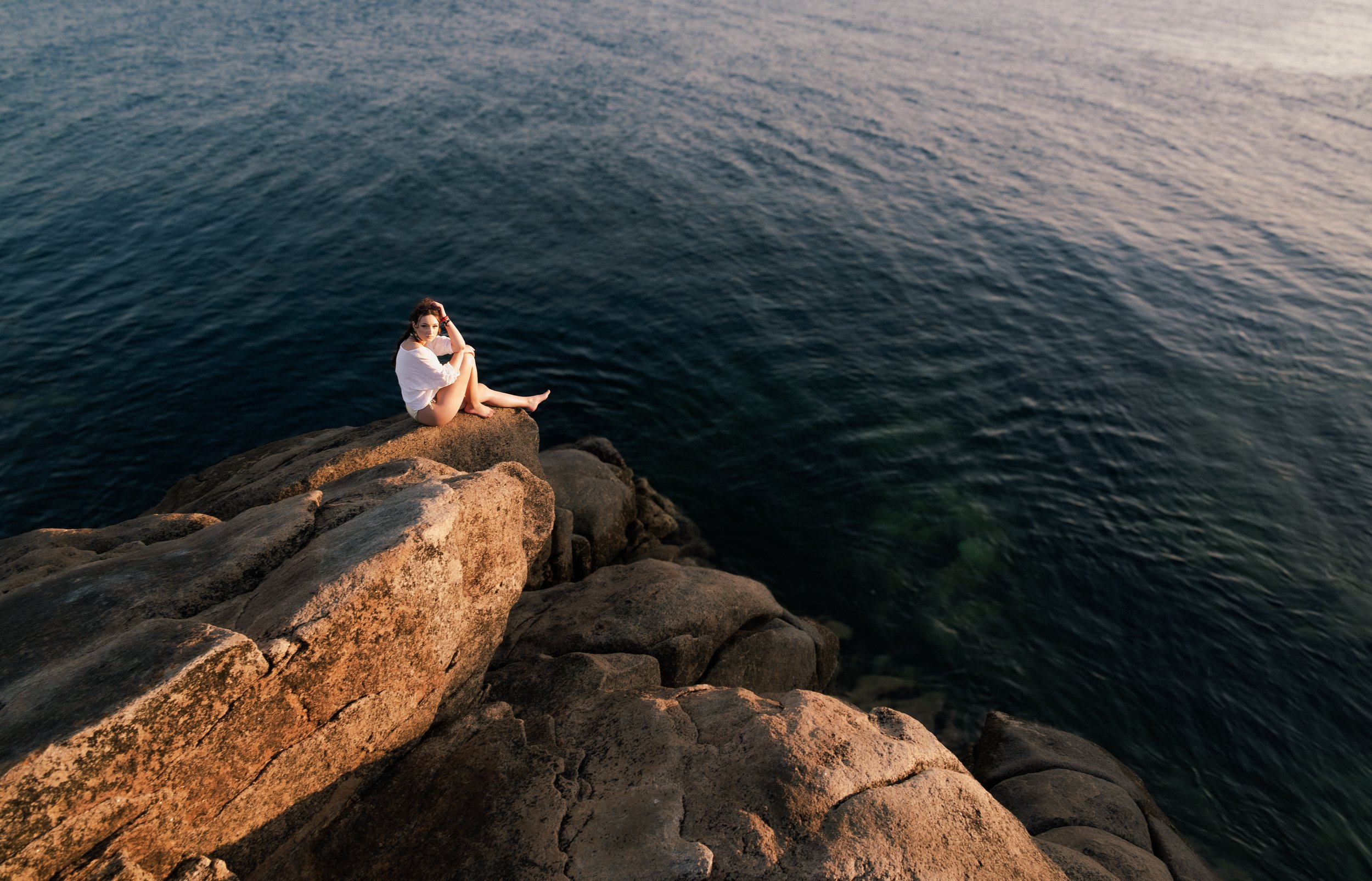 Jeune femme assise sur un rocher au bord de la mer, regardant l'eau, lors du coucher de soleil.
