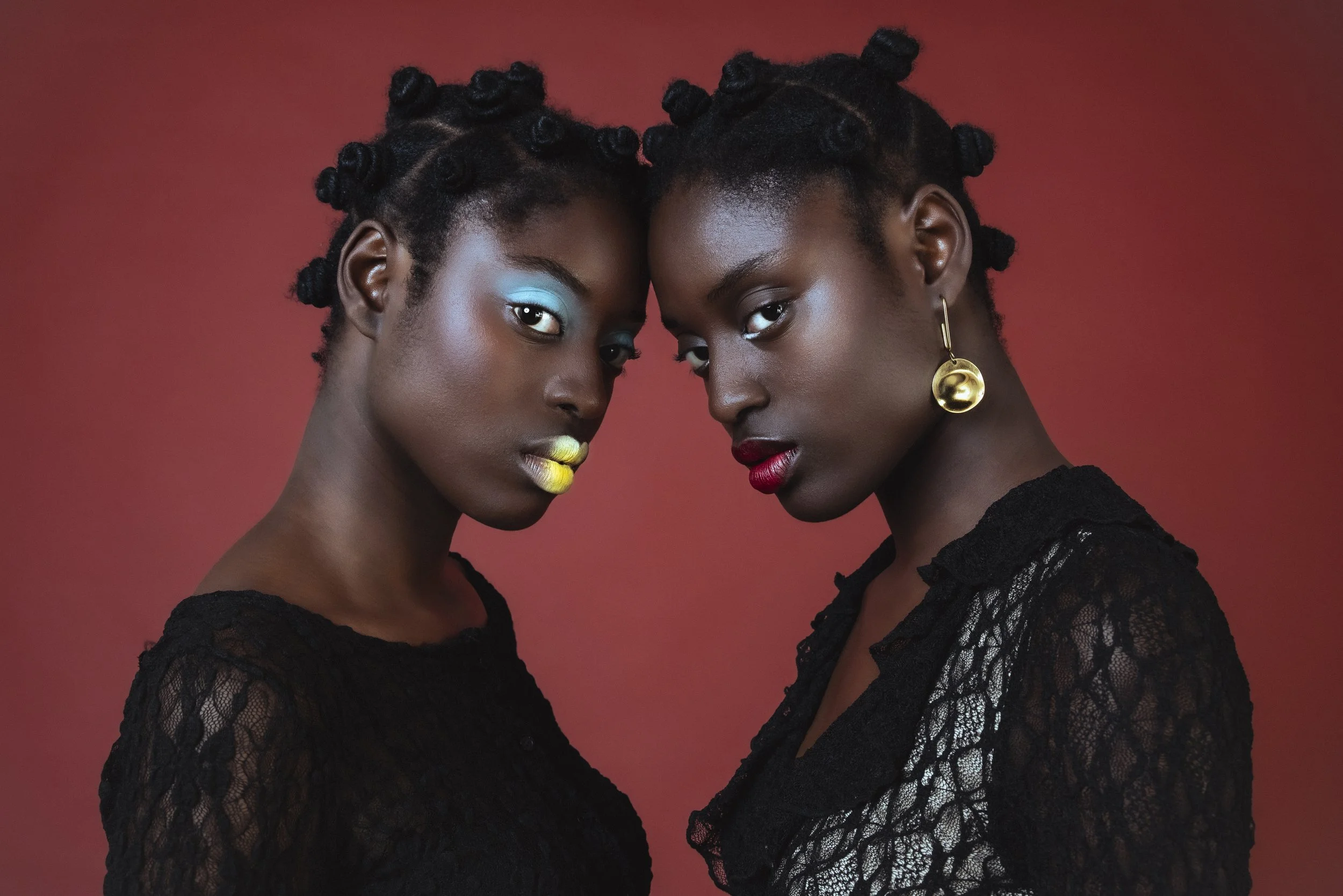 Deux femmes noires avec des coiffures bantu, portant des vêtements en dentelle noire, posant contre un fond rouge, avec un maquillage coloré et des bijoux dorés.