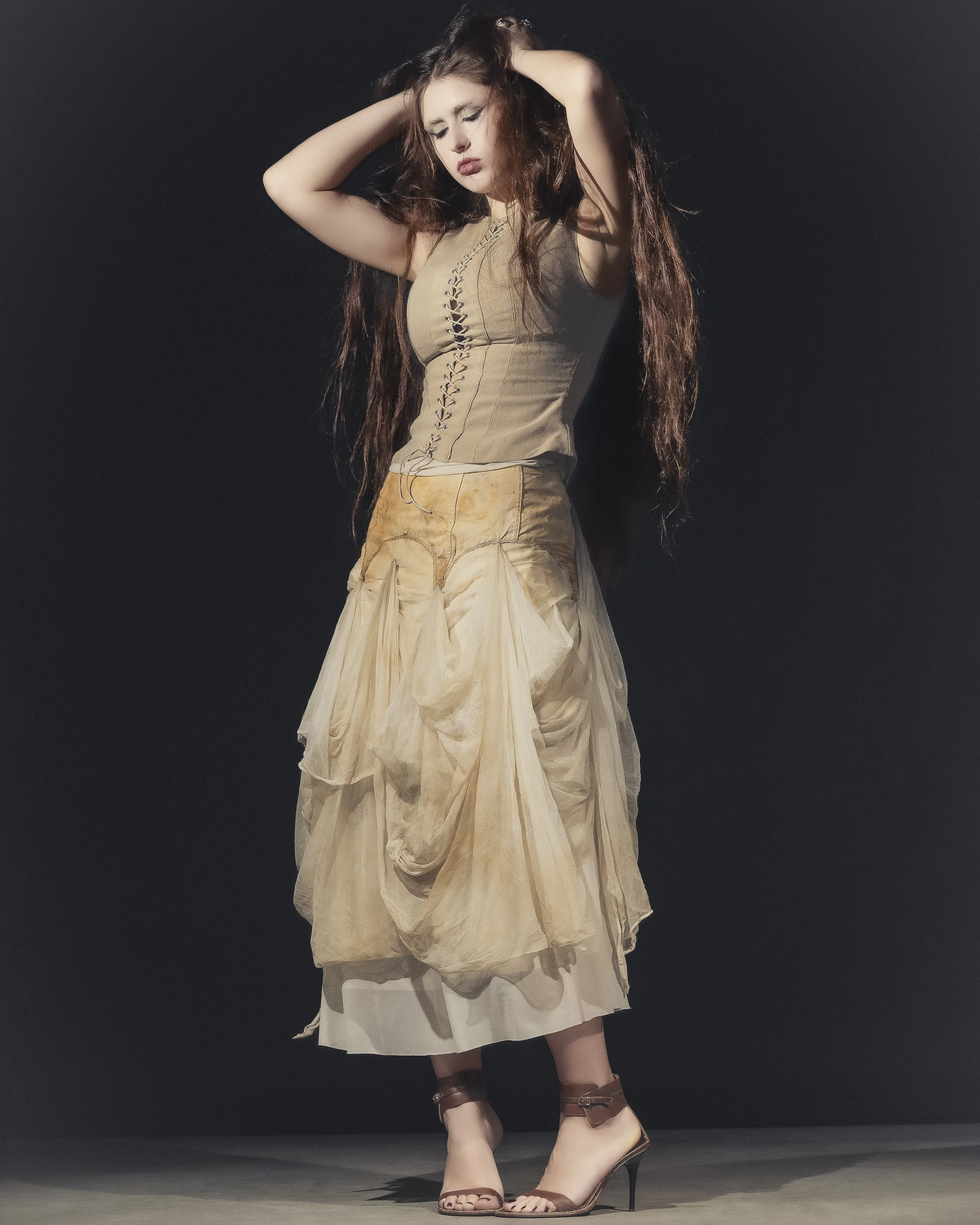 Une femme posant devant un fond noir, portant une robe beige avec des détails en tissu léger et des sandales à talons.