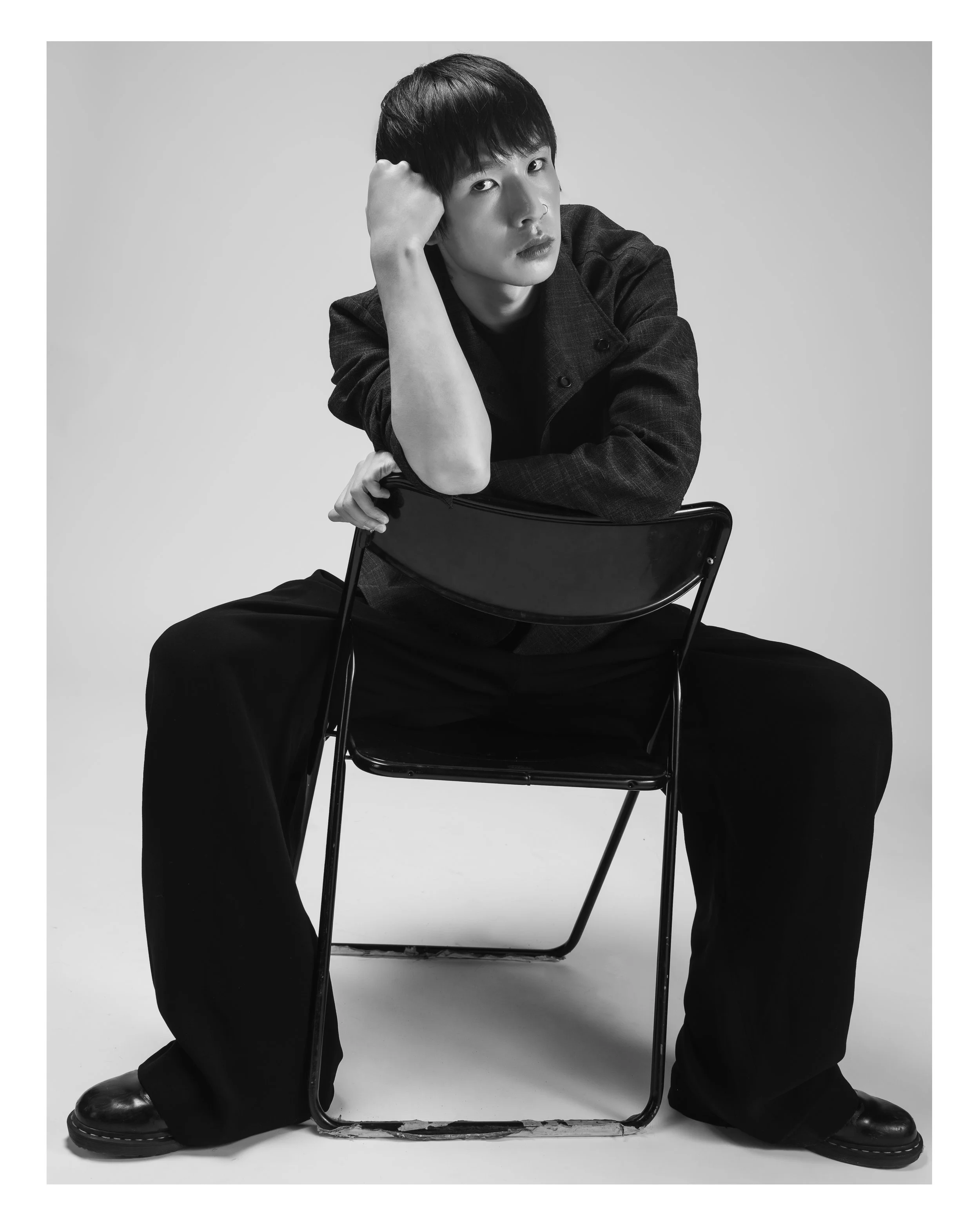 Une personne assise sur une chaise, en noir et blanc, avec une expression sérieuse, portant une veste foncée et des pantalons amples, les jambes écartées et la tête reposant sur la main, dans un studio avec fond clair.