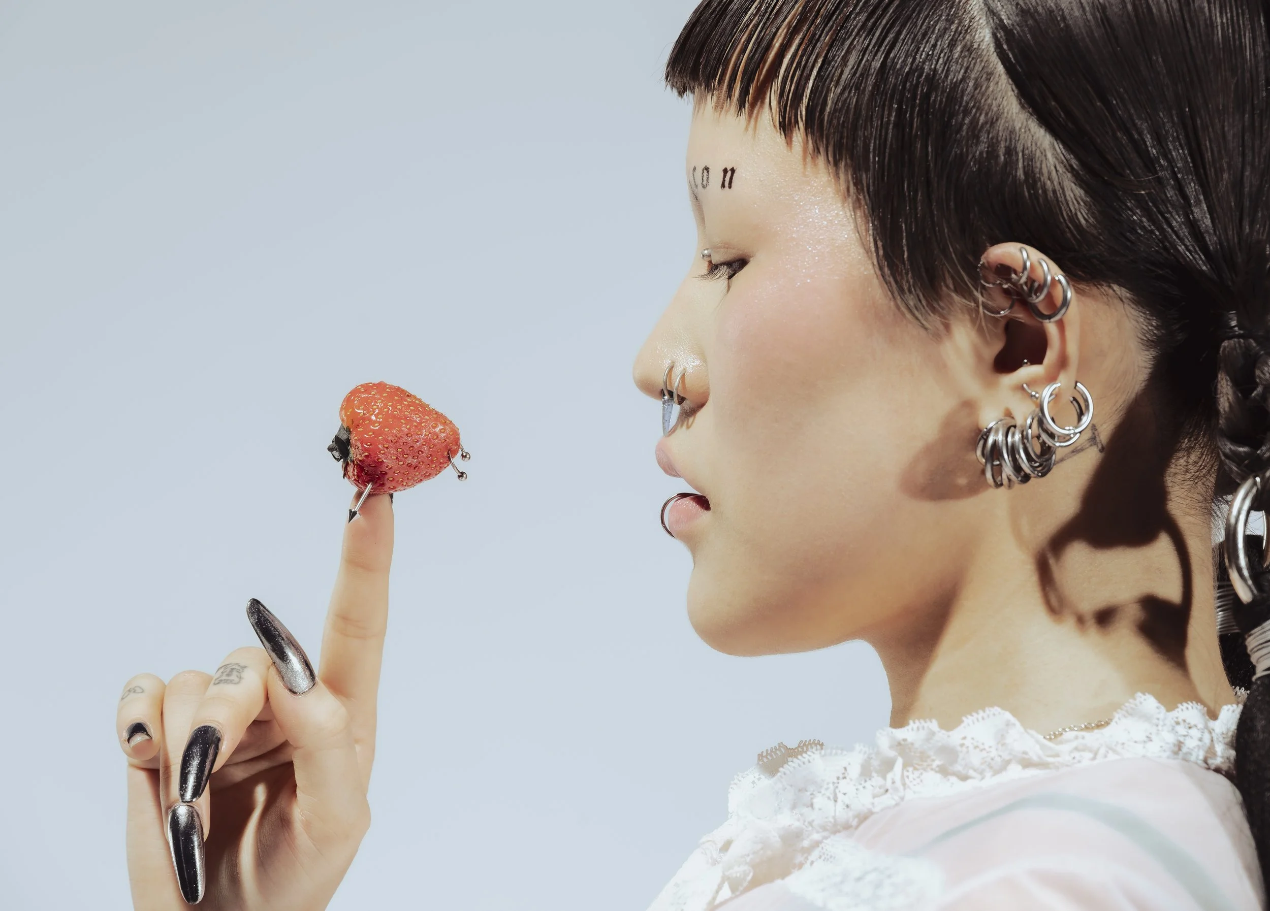 Une jeune femme aux nombreux piercings, avec des cheveux courts et bruns, tient un doigt avec un doigté à la place du bout, sur lequel repose une fraise, contre un fond bleu clair.
