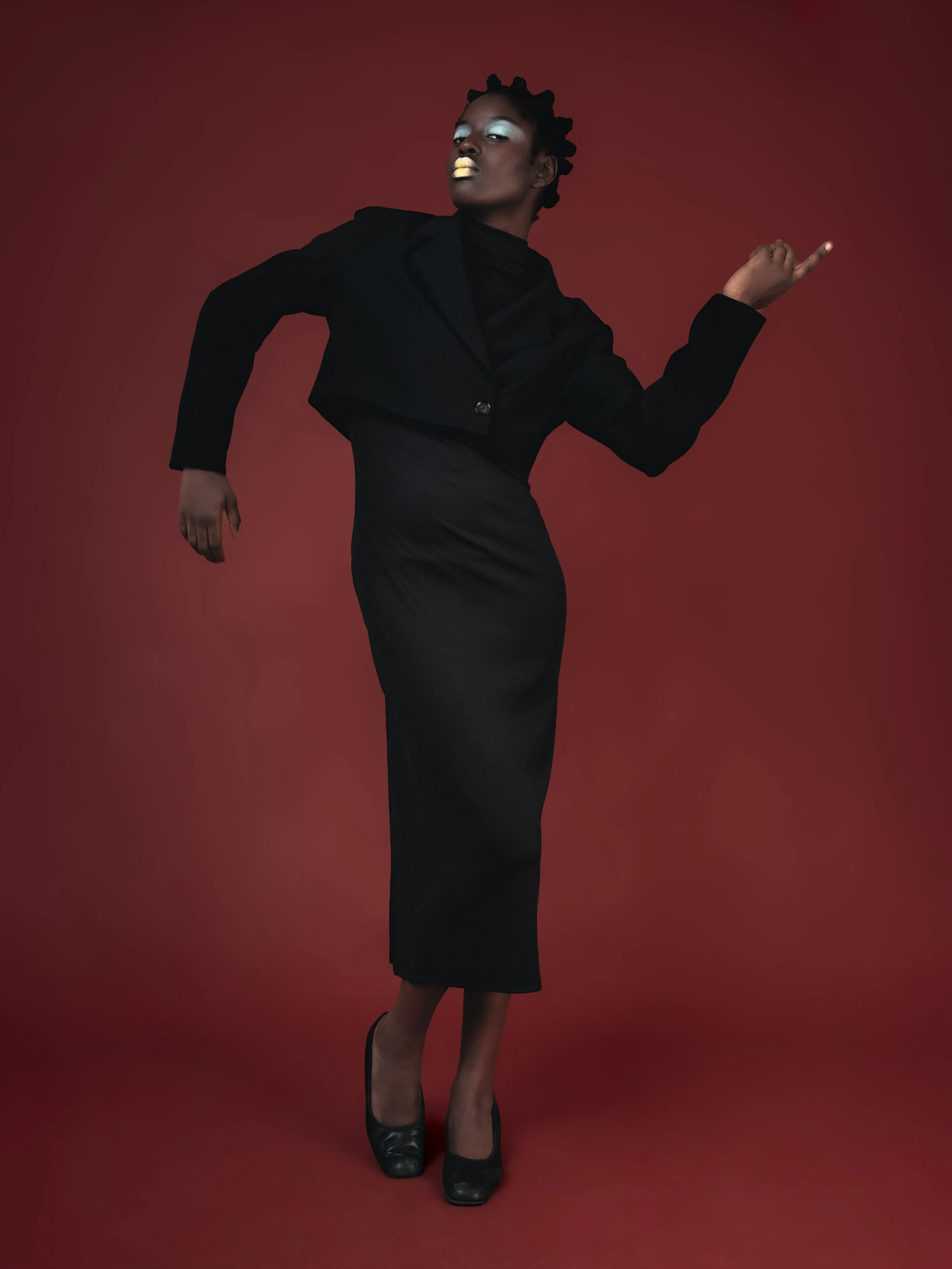 Une femme élégante en tenue noire, portant des talons noirs, se tient contre un fond rouge, avec une pose confiante et sophistiquée.