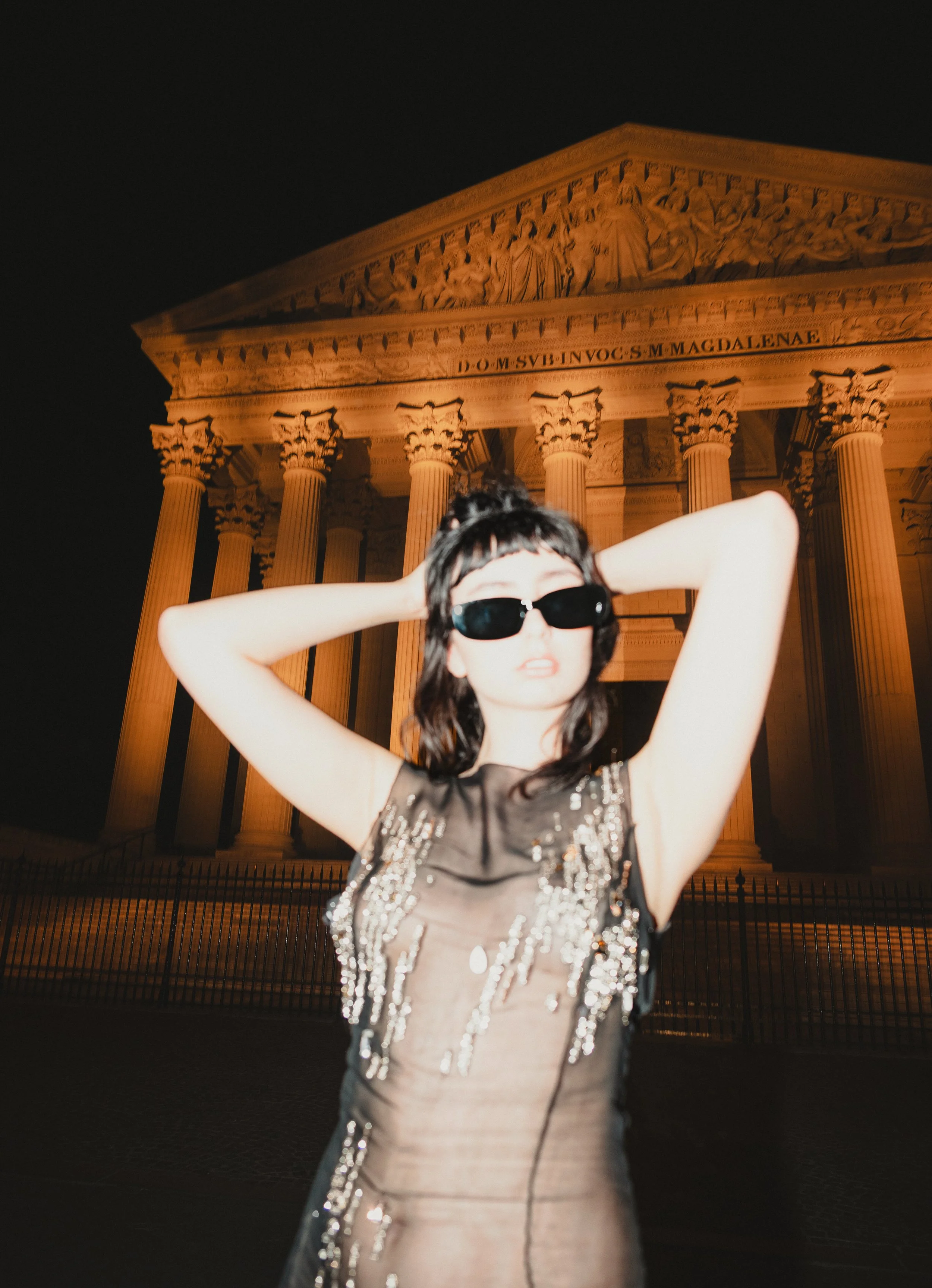 Une jeune femme portant des lunettes de soleil noires et une robe brillante à sequins se tient devant le Panthéon à Paris, la nuit.