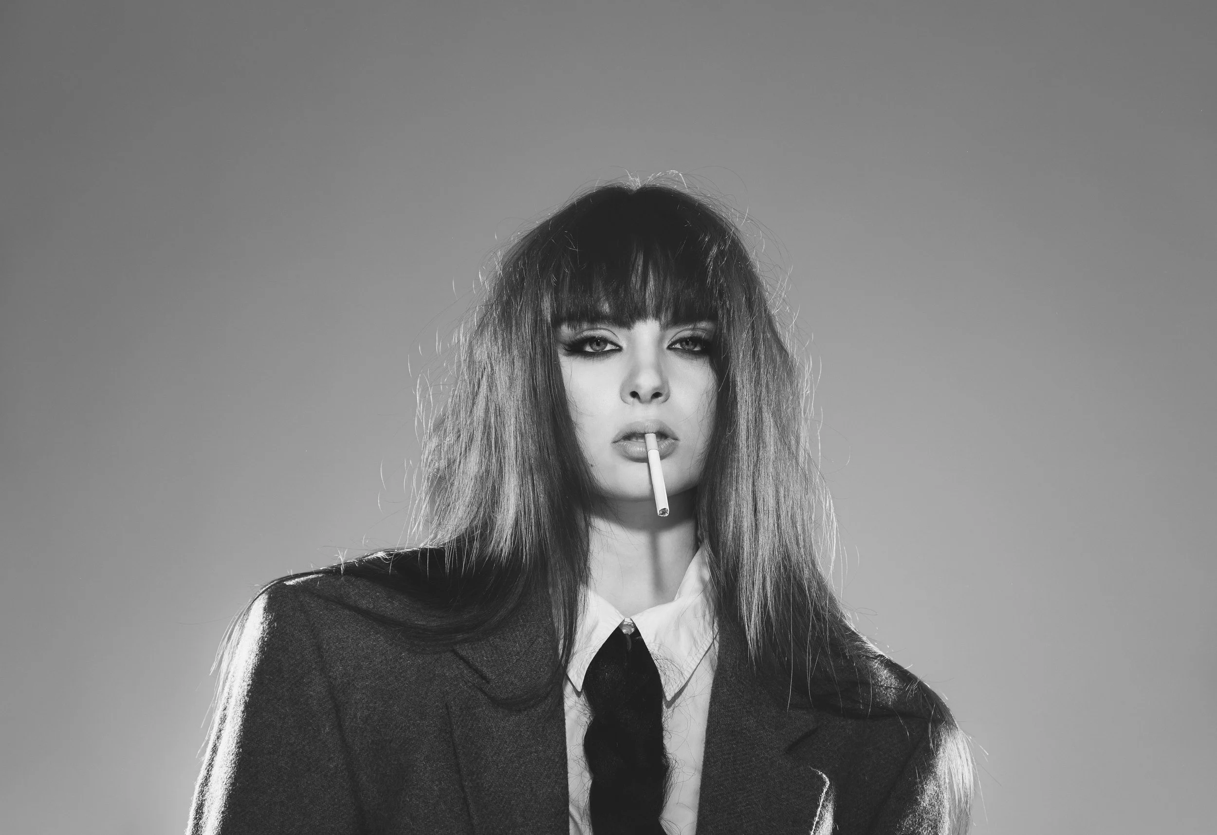 Une femme avec des cheveux longs et volumineux, portant une veste en tissu et une chemise avec un nœud noir, tenant une cigarette entre les lèvres, dans un style noir et blanc.