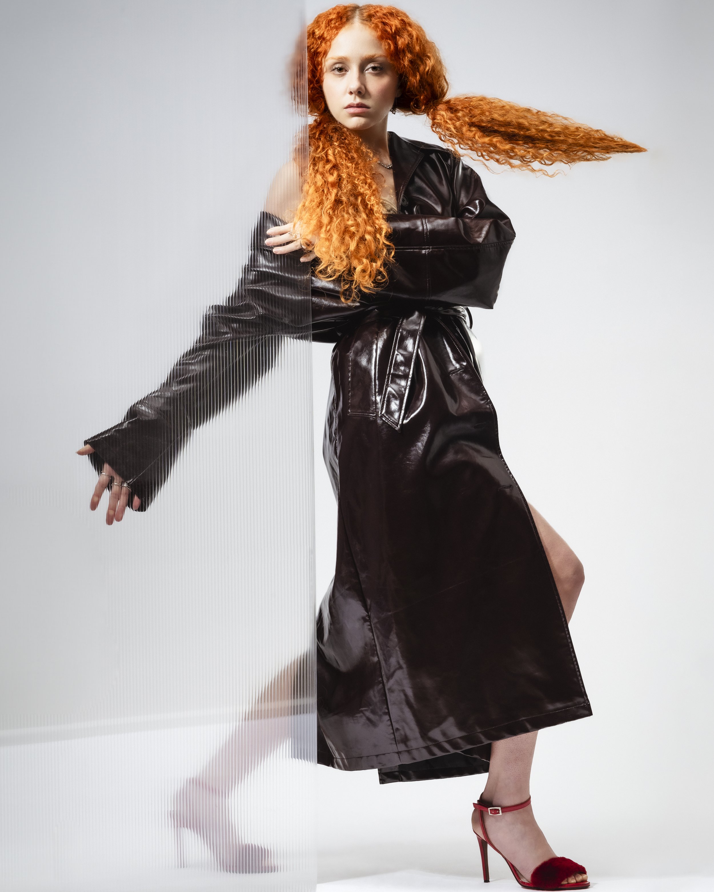 Femme avec cheveux roux, portant une robe en cuir noire et des chaussures à talons rouges, posant contre un mur partiellement transparent.