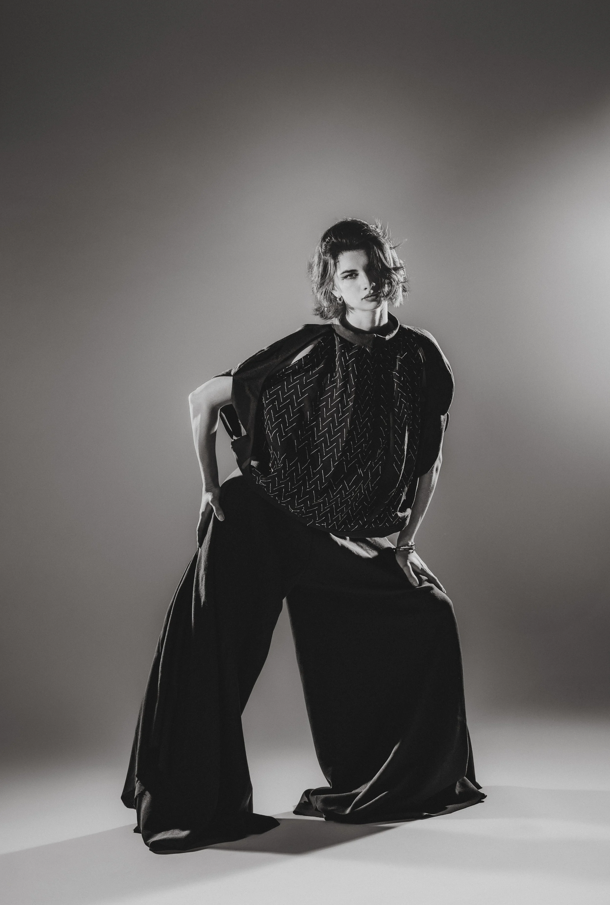 Une femme pose en tenue noire dans un studio avec un éclairage contrasté, regardant vers l'objectif.