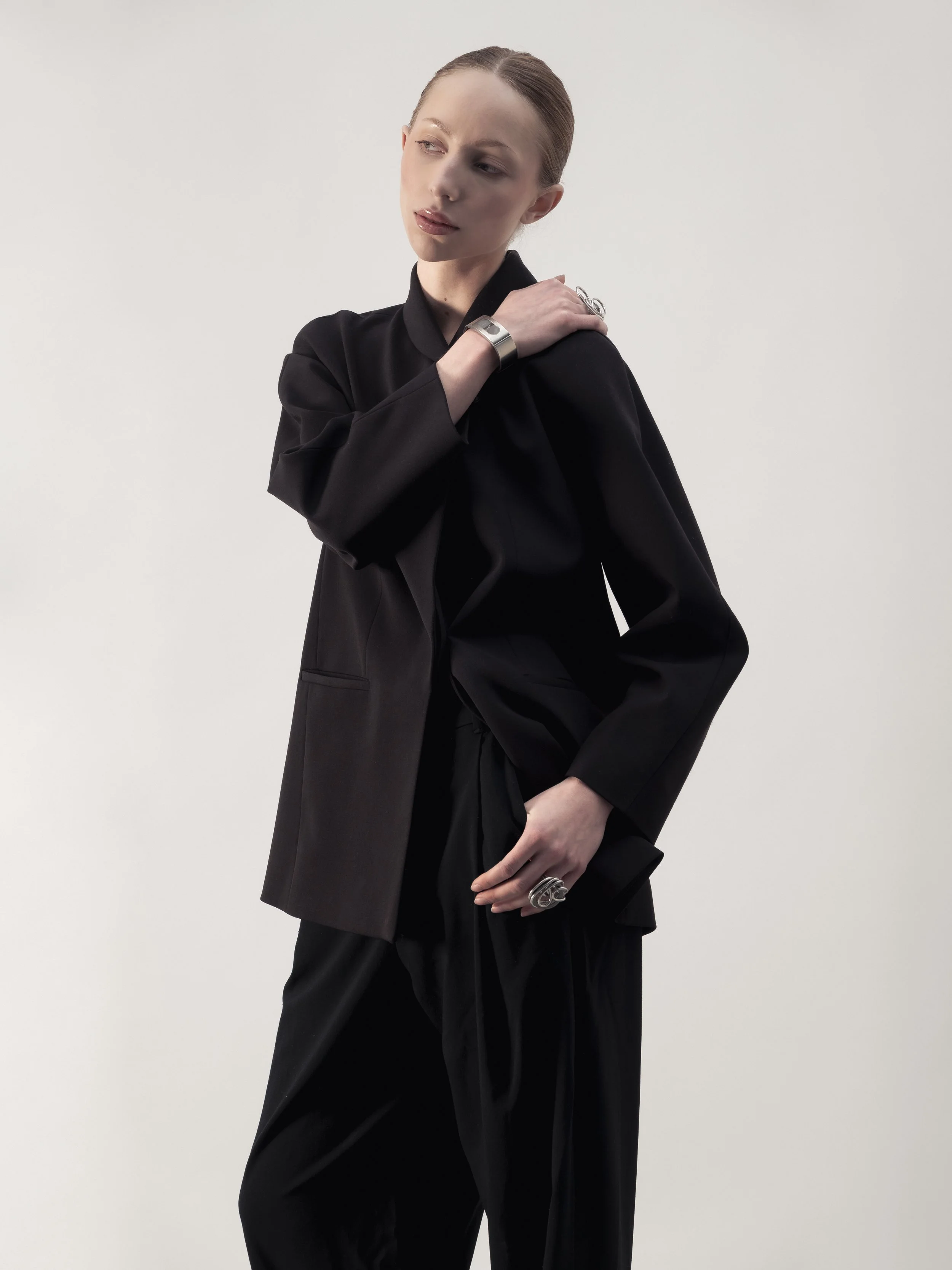 Une femme portant un tailleur noir avec des bijoux en argent, posant contre un fond blanc.