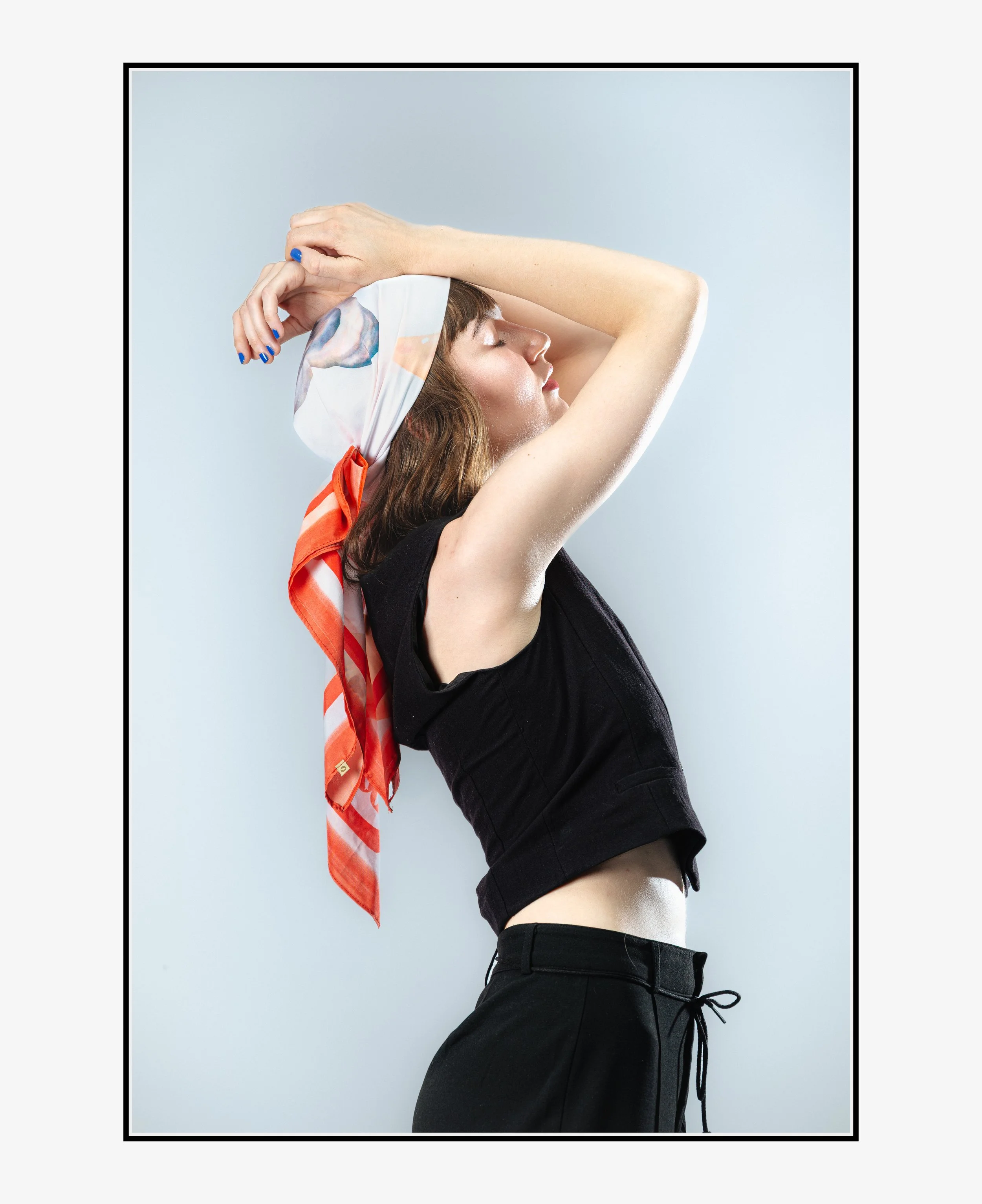 Une femme pose avec ses yeux fermés, portant un bandeau coloré et un haut noir sans manches, les bras levés au-dessus de sa tête contre un mur blanc.