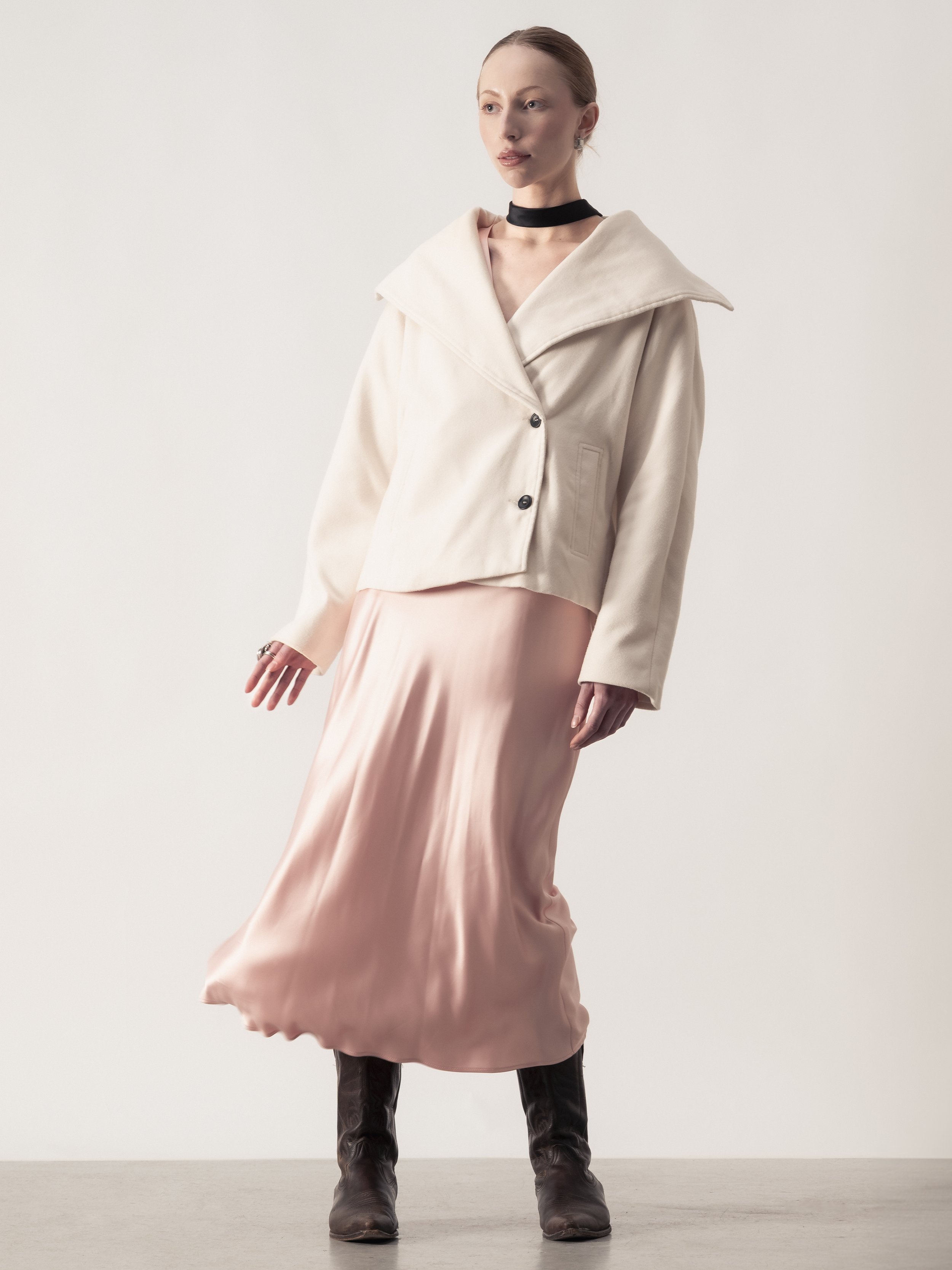 Une femme portant une veste beige, une jupe longue en satin rose, des bottes en cuir marron, un collier noir et des boucles d'oreilles, posant devant un fond blanc.
