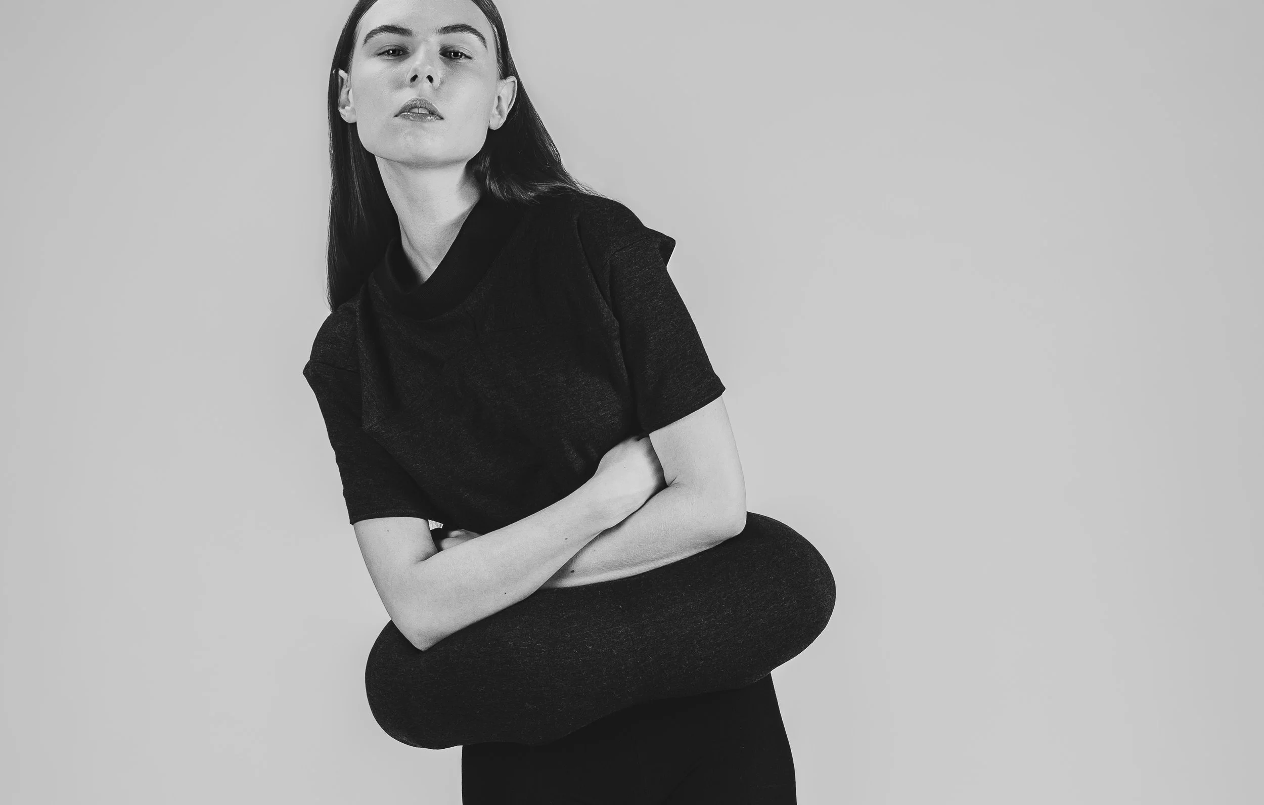 Une femme en noir et blanc, portant un t-shirt noir, tenant un vêtement contre sa poitrine, et posant contre un fond neutre.