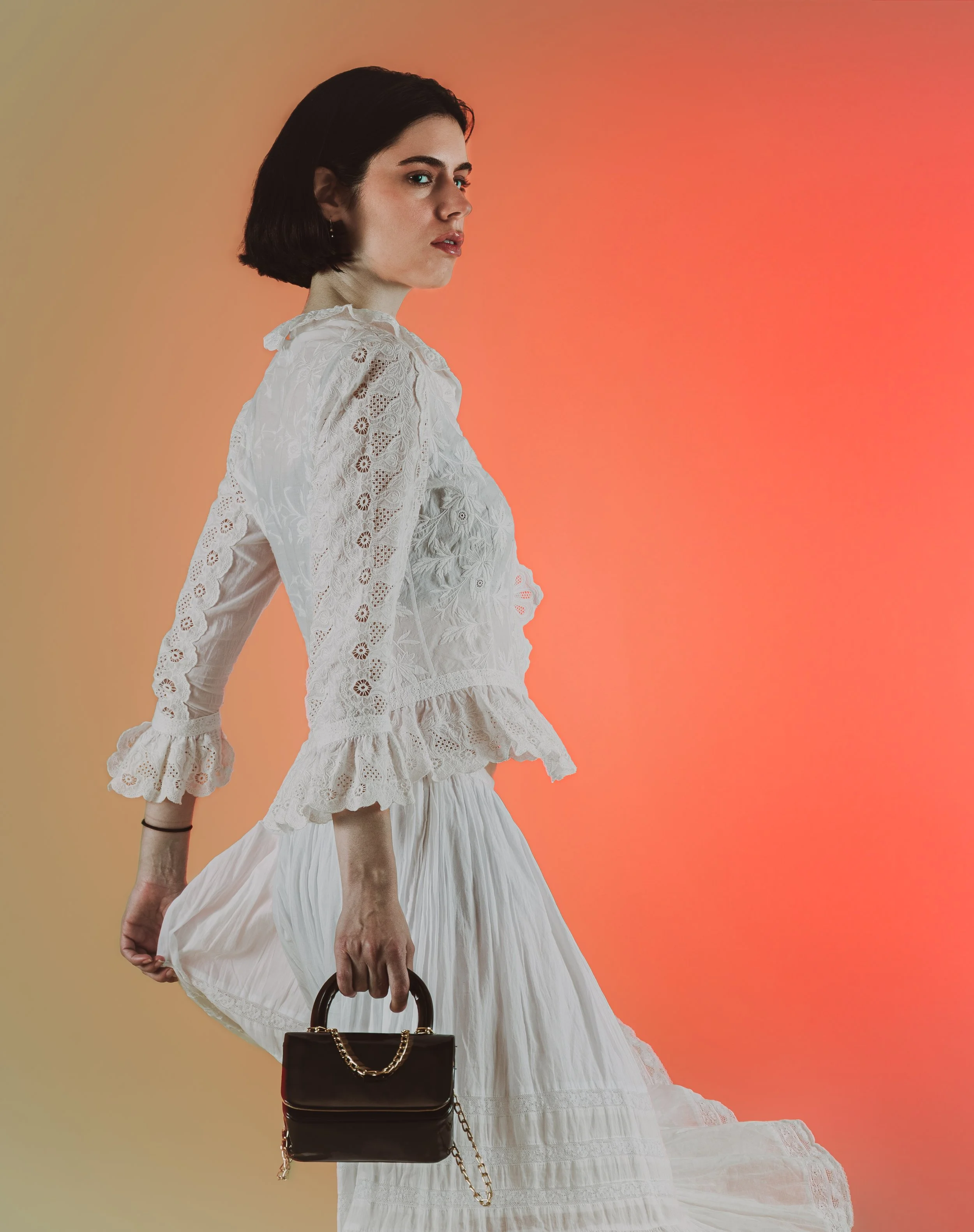 Une femme portant une robe en dentelle blanche, tenant un sac à main noir avec une chaîne en or, devant un fond dégradé de couleurs chaudes.