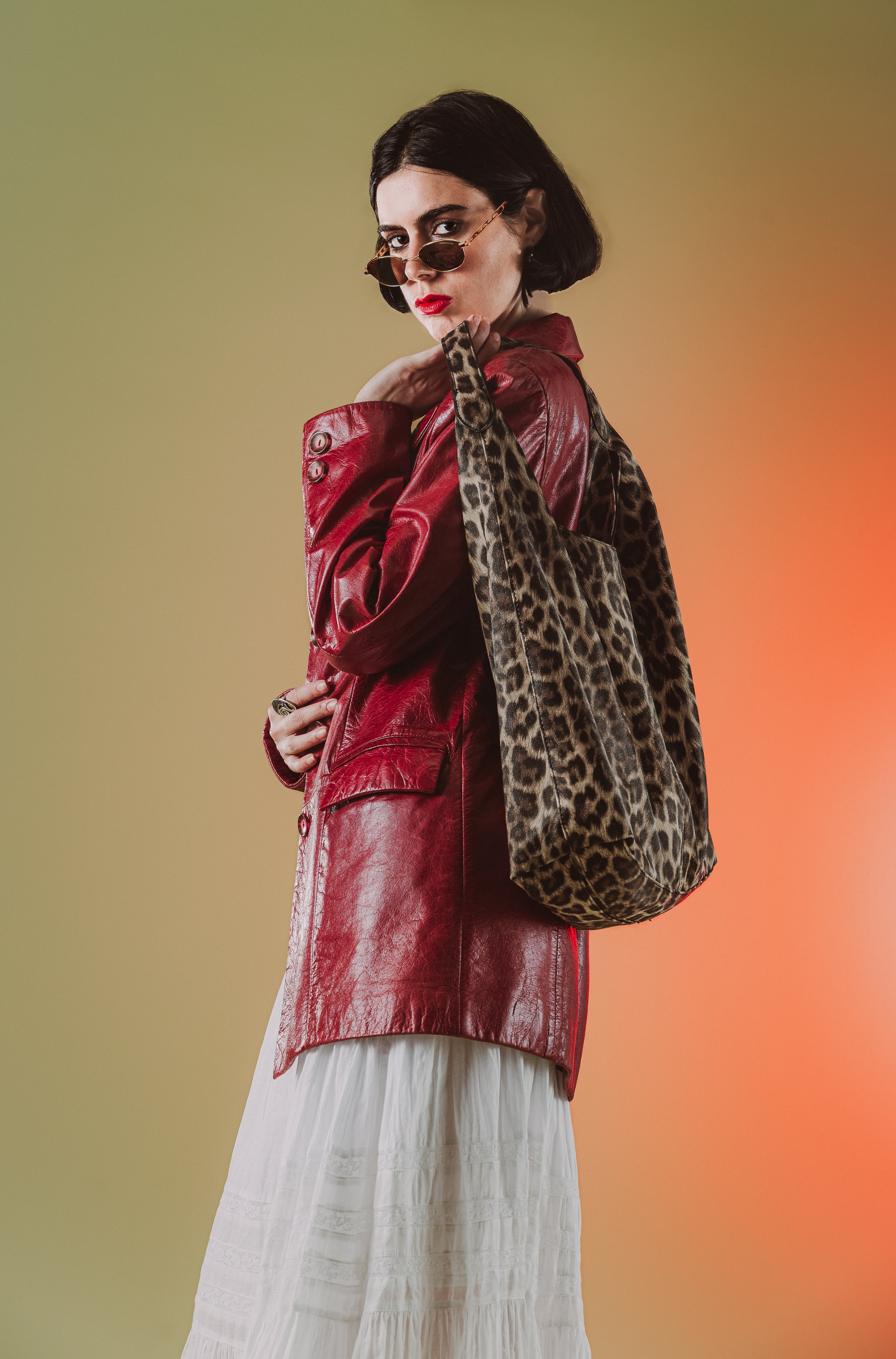 Une femme avec des cheveux noirs courts, portant des lunettes de soleil, une veste en cuir rouge, une jupe blanche et tenant un sac à main à motif léopard, pose devant un fond coloré dégradé allant du vert à l'orange.