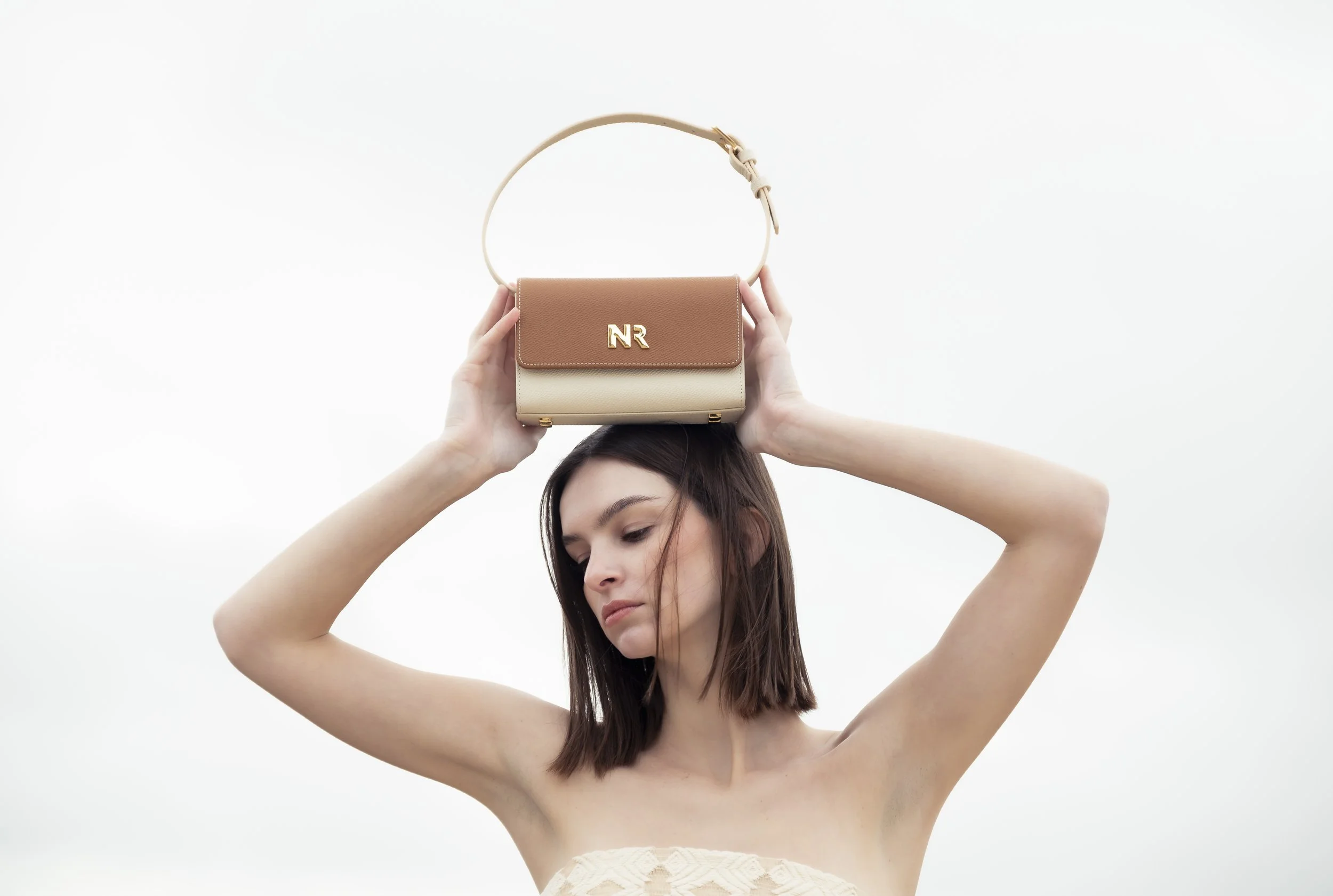 Femme tenant un sac à main beige et marron au-dessus de la tête, vêtements sans manches, cheveux bruns courts, fond blanc.