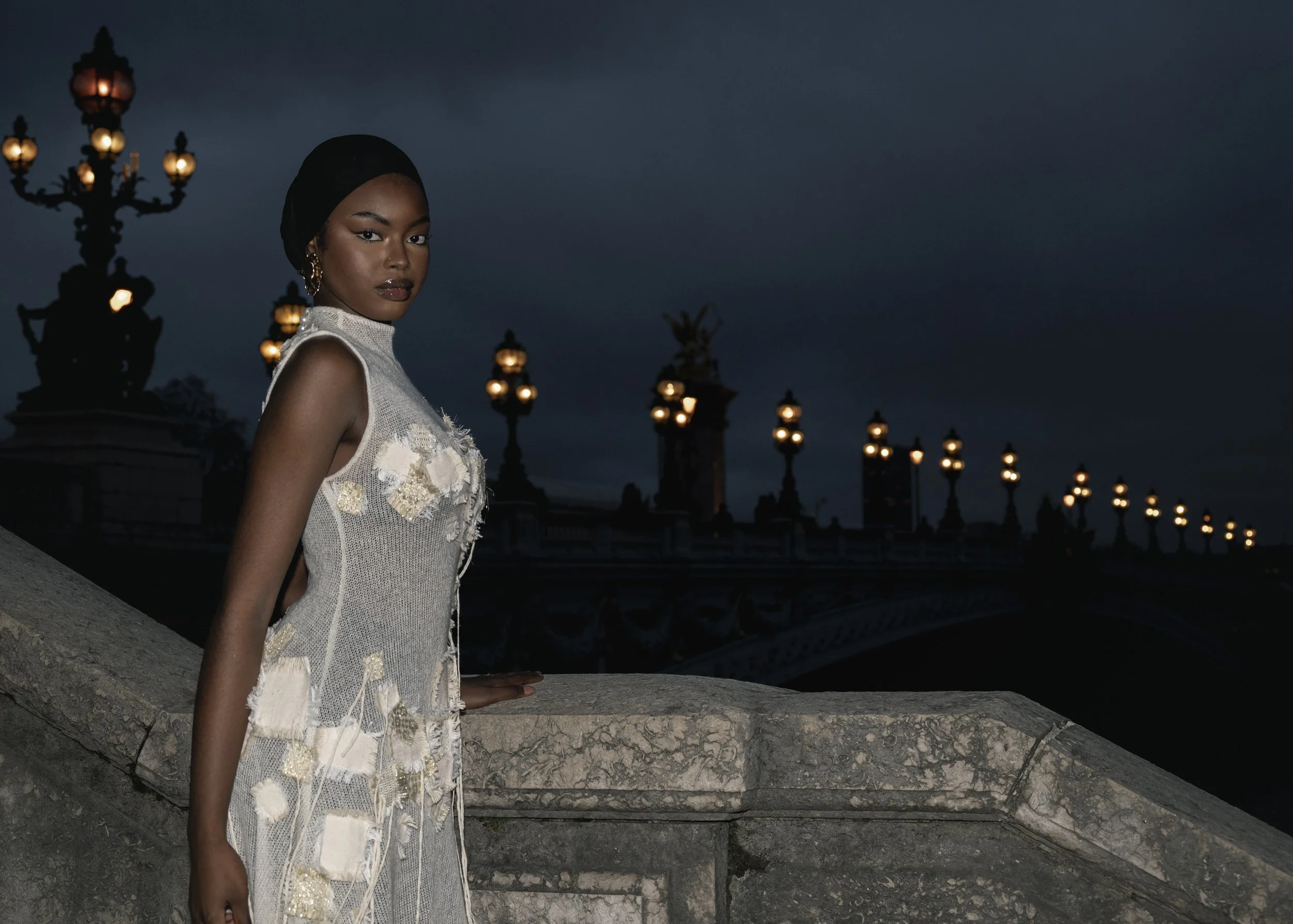 Une femme noire pose sur un pont la nuit, portant une robe élégante avec des détails en relief, avec des lampadaires en arrière-plan.