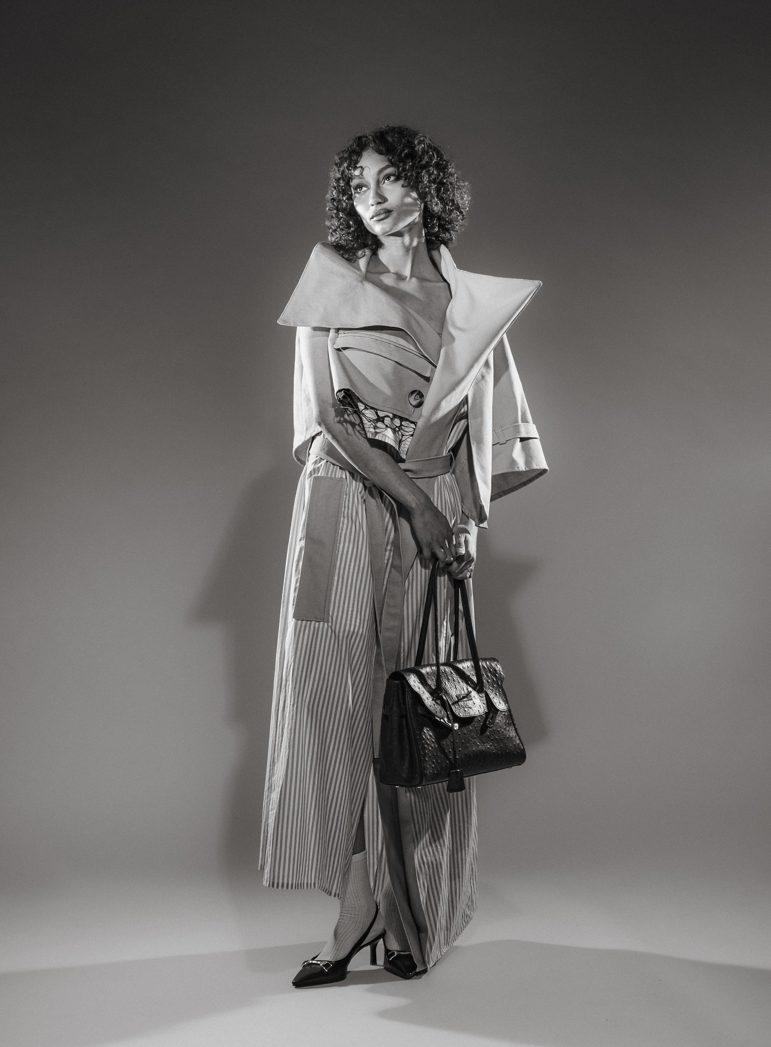 Une femme porte un trench beige, un pantalon large à rayures et des chaussures à talons, elle tient un sac à main en cuir noir, devant un fond uni en couleur sombre.