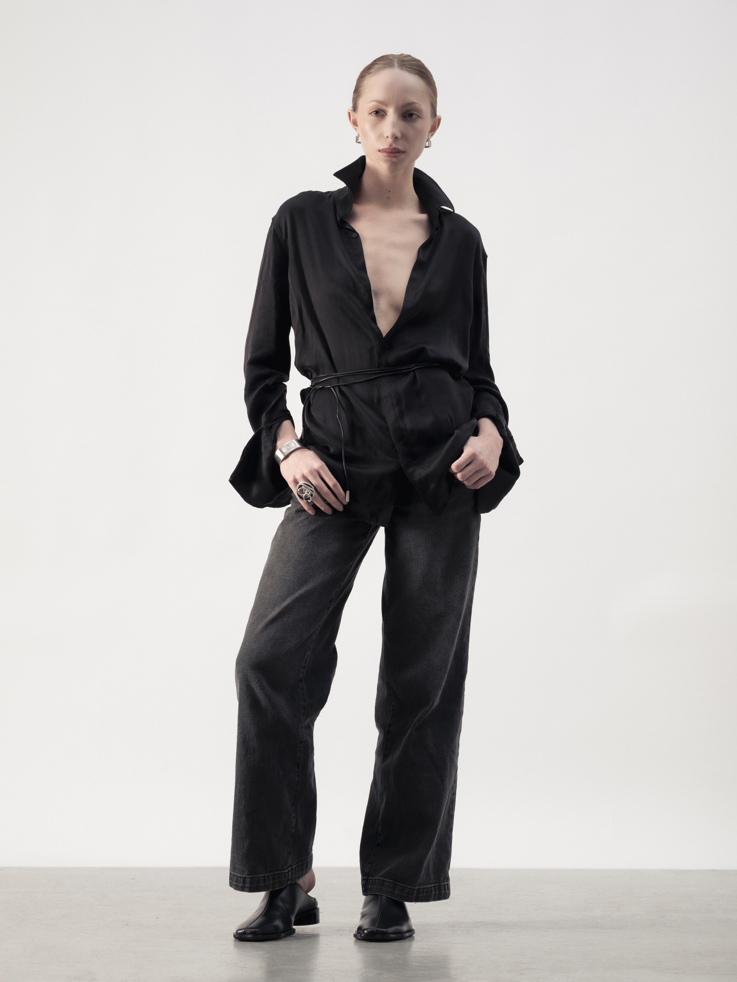 Femme en vêtements noirs, pantalon large, blouse ouverte, debout sur un fond blanc.