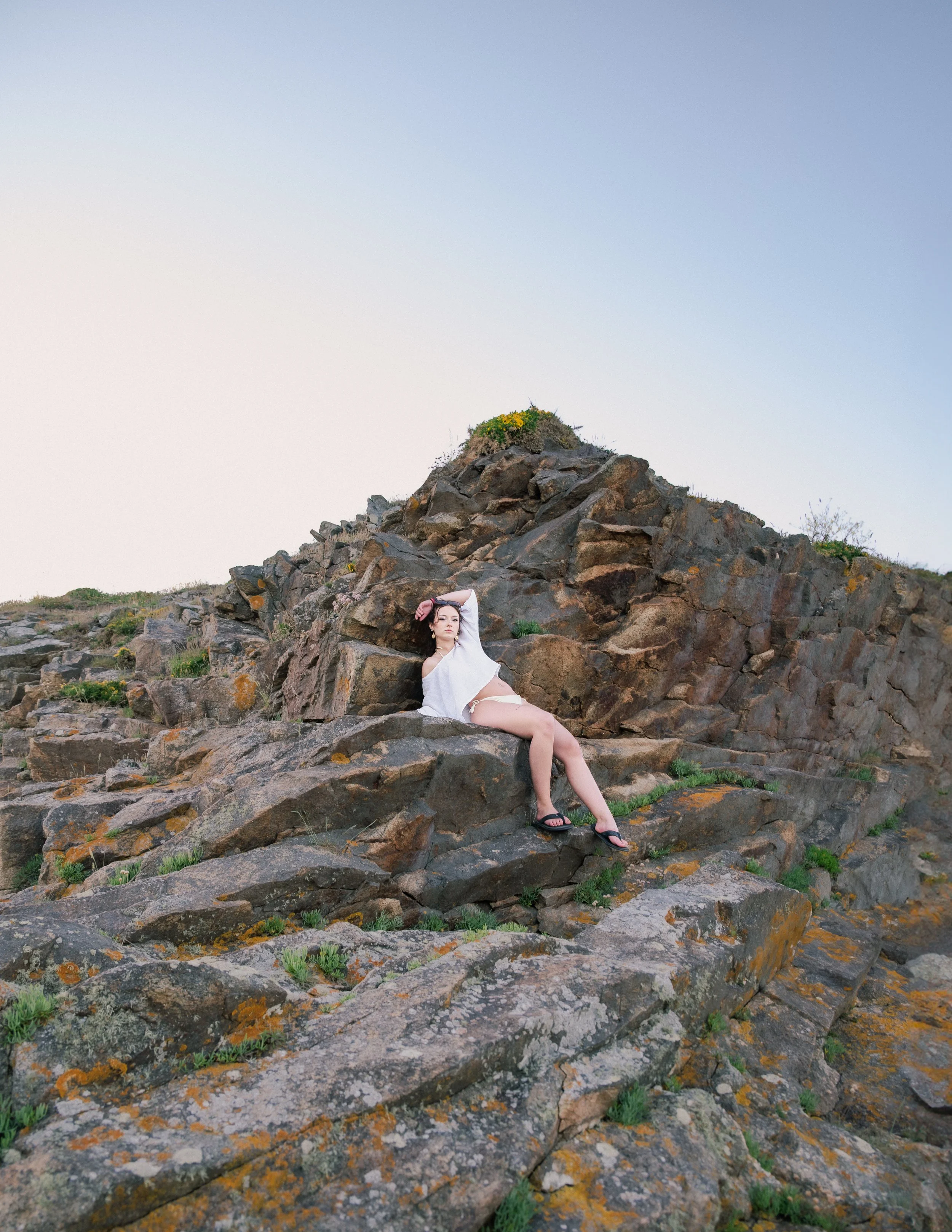 Femme en maillot blanc et foulard noir assise sur des rochers en pleine nature, avec un ciel clair en arrière-plan.
