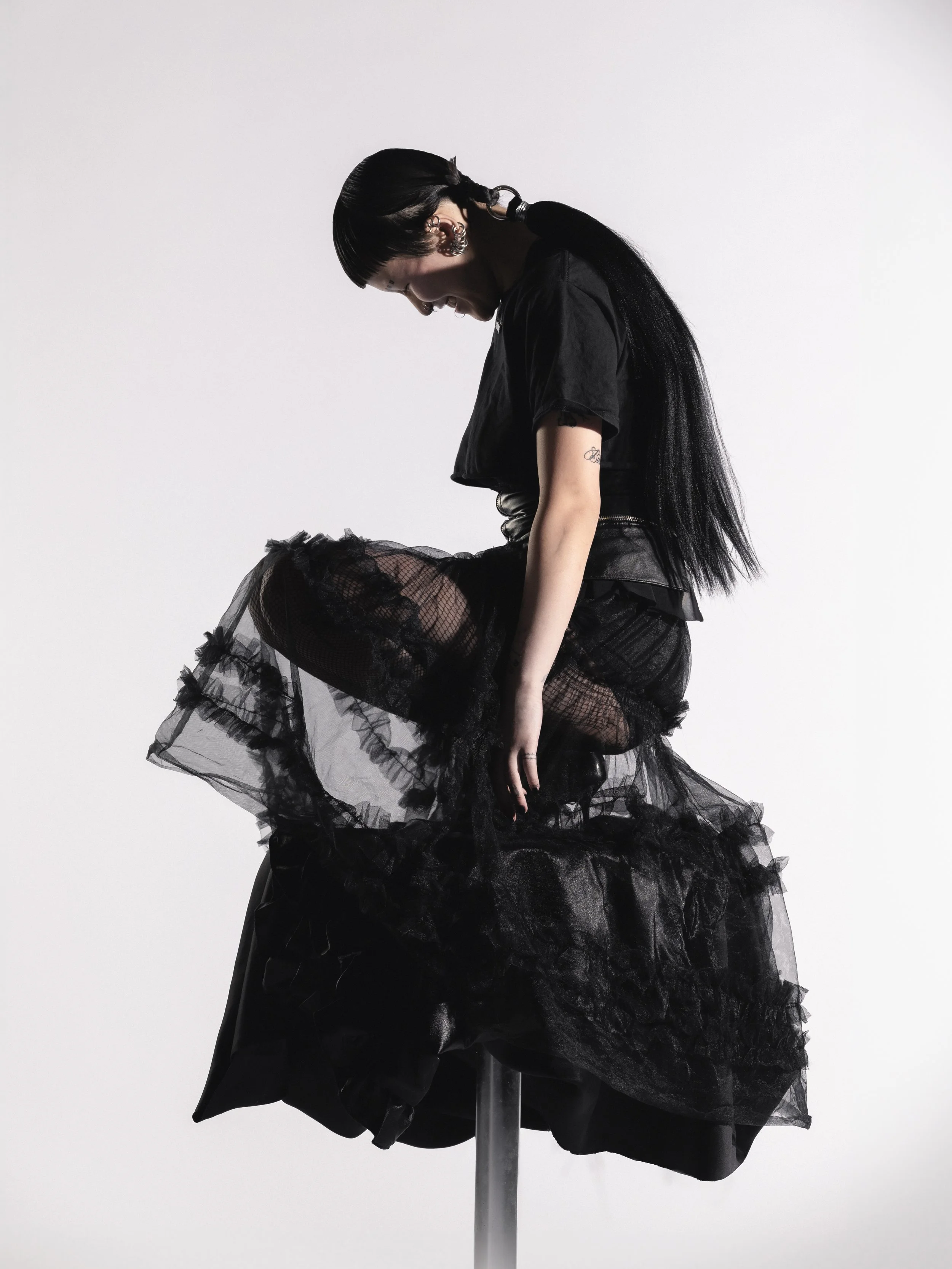 Femme vêtue d'une robe noire en tulle, assise sur une plateforme blanche contre un fond blanc, regardant vers le bas, avec style gothique. 