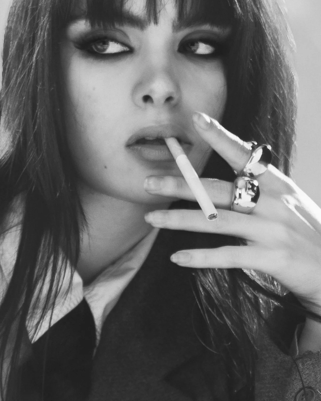 Portrait en noir et blanc d'une femme avec maquillage marqué, tenant une cigarette entre les doigts, portant plusieurs bagues.