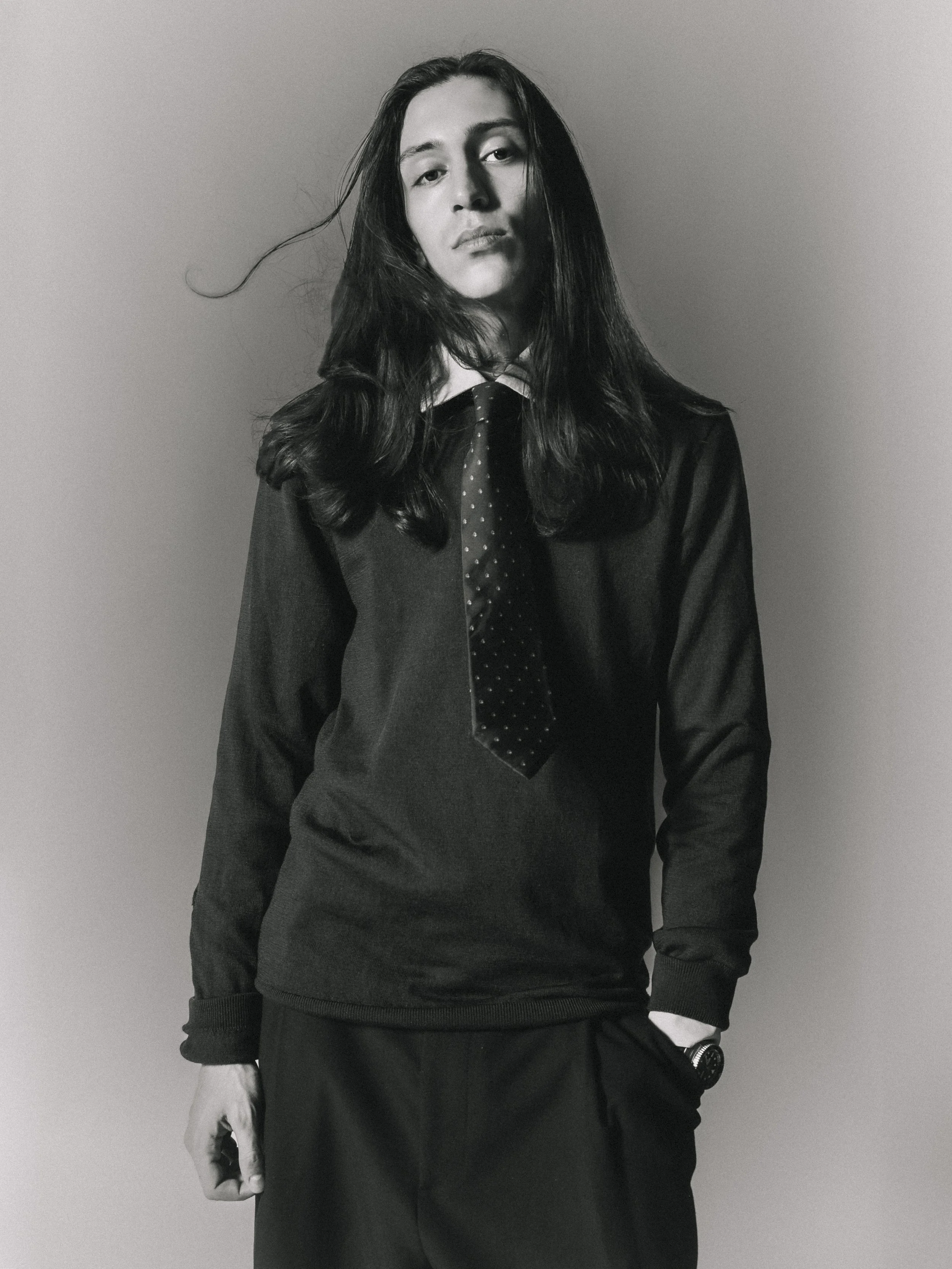 Une femme en vêtement sombre, vêtue d'une chemise et d'une veste, avec une cravate à pois, pose contre un mur gris en noir et blanc.