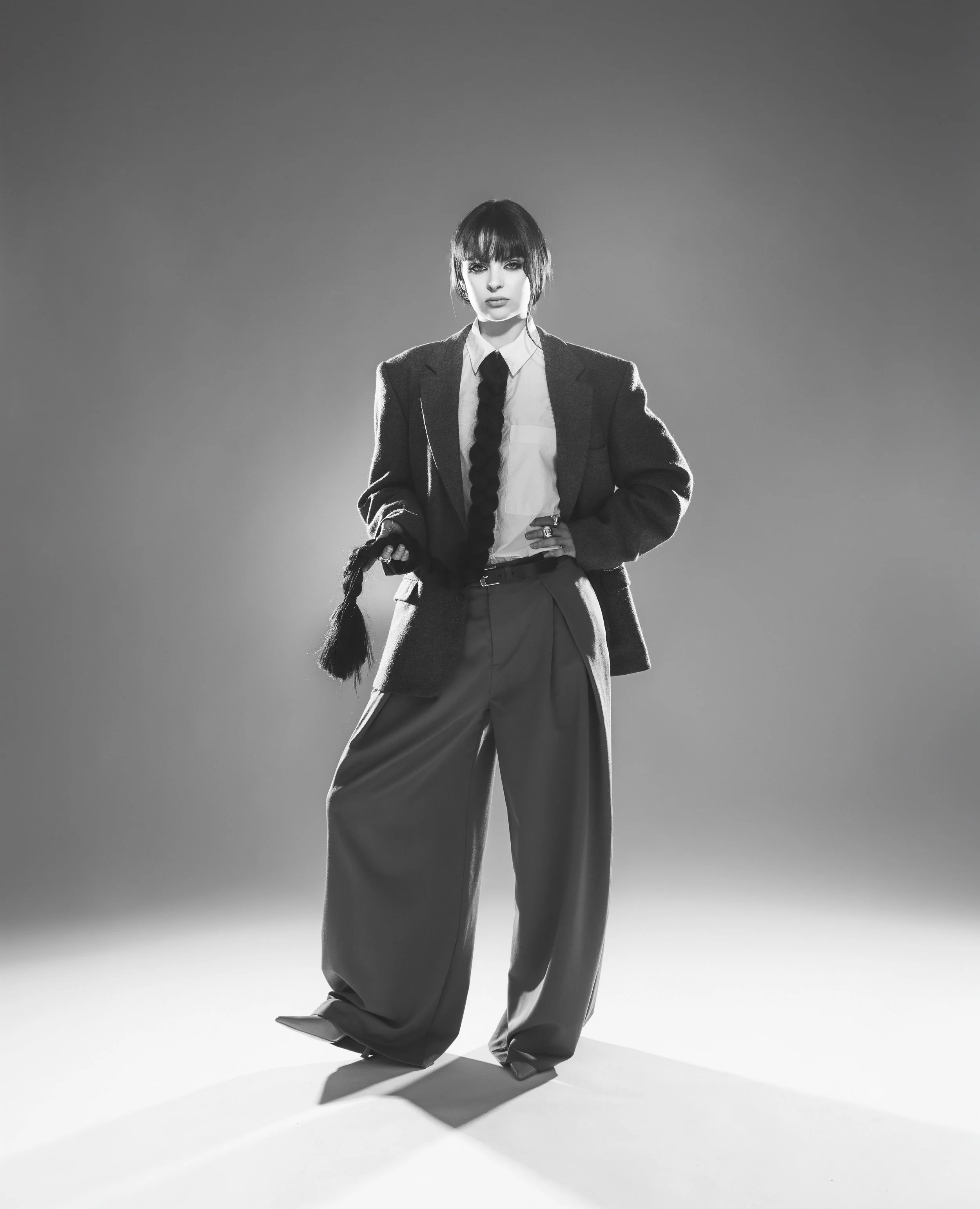 Une femme posa en tenue formelle avec un blazer, une chemise blanche, un pantalon ample, dans un studio avec lumière modulée, en noir et blanc.