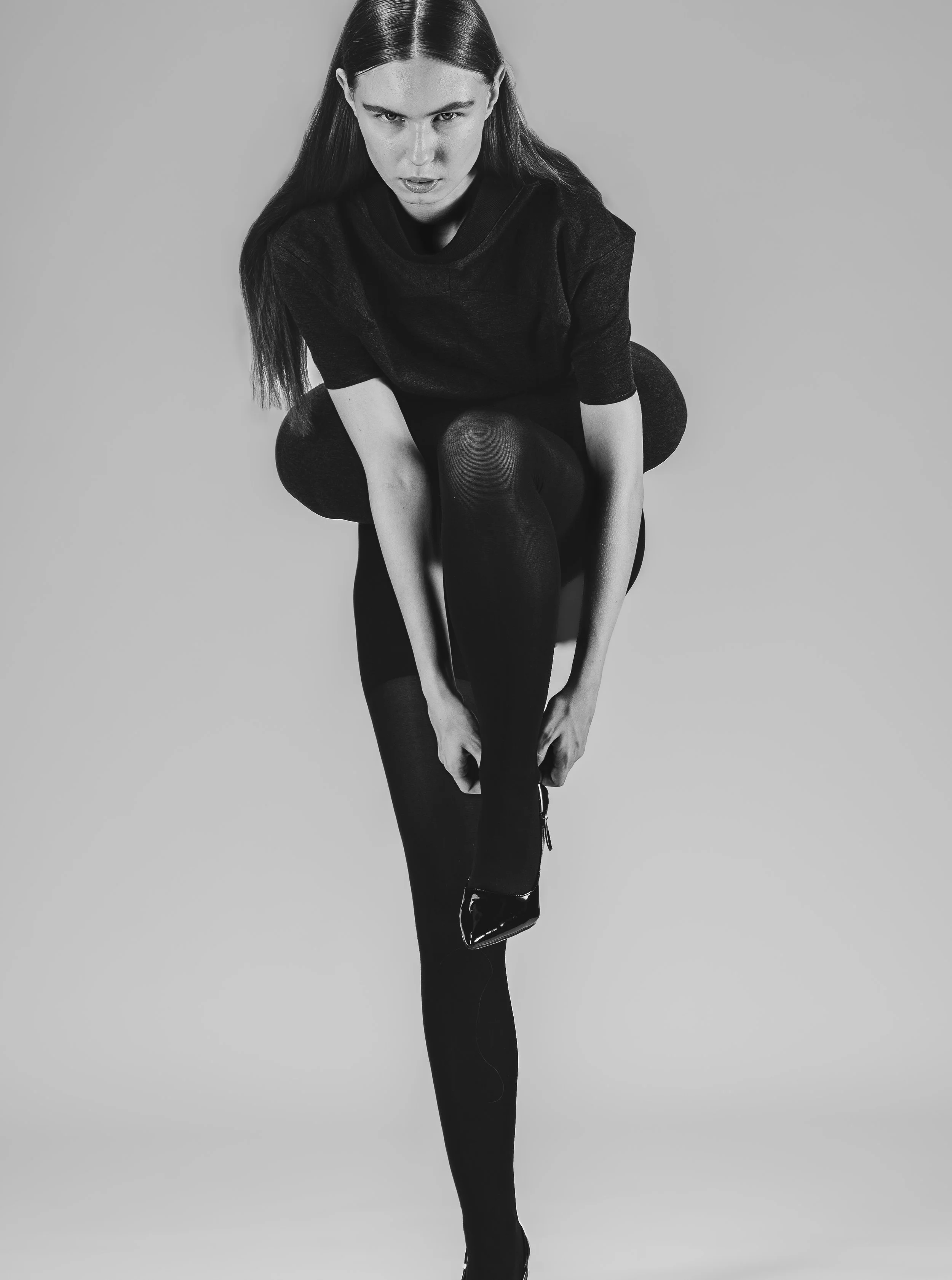 Femme en noir et blanc, assise en position de yoga ou d'étirement, regardant intensément vers l'avant, avec le visage légèrement incliné, portant des vêtements sombres et des collants.