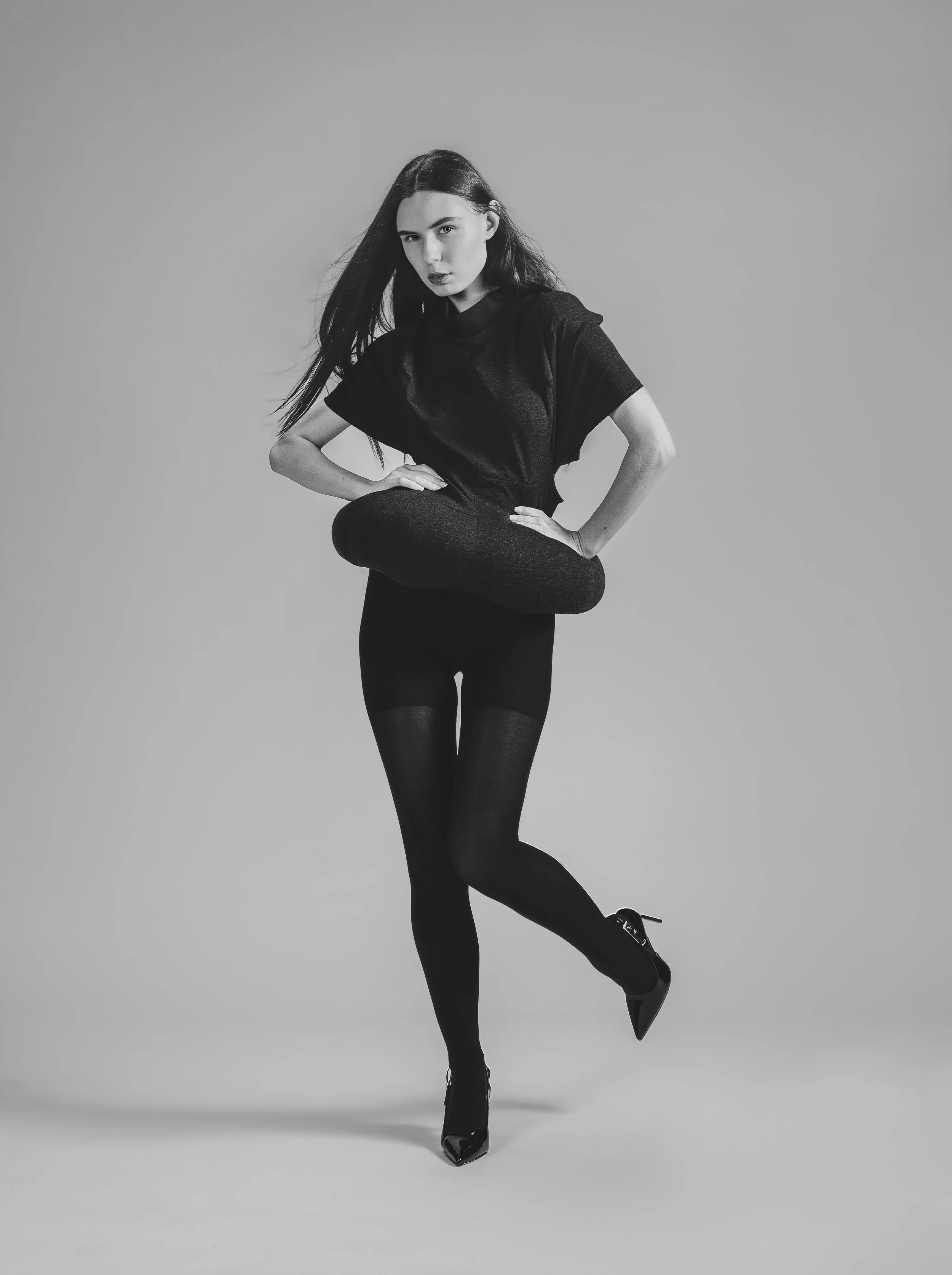 Femme portant un légging noir et une chemise noire, posant avec une jambe levée, dans une image en noir et blanc.