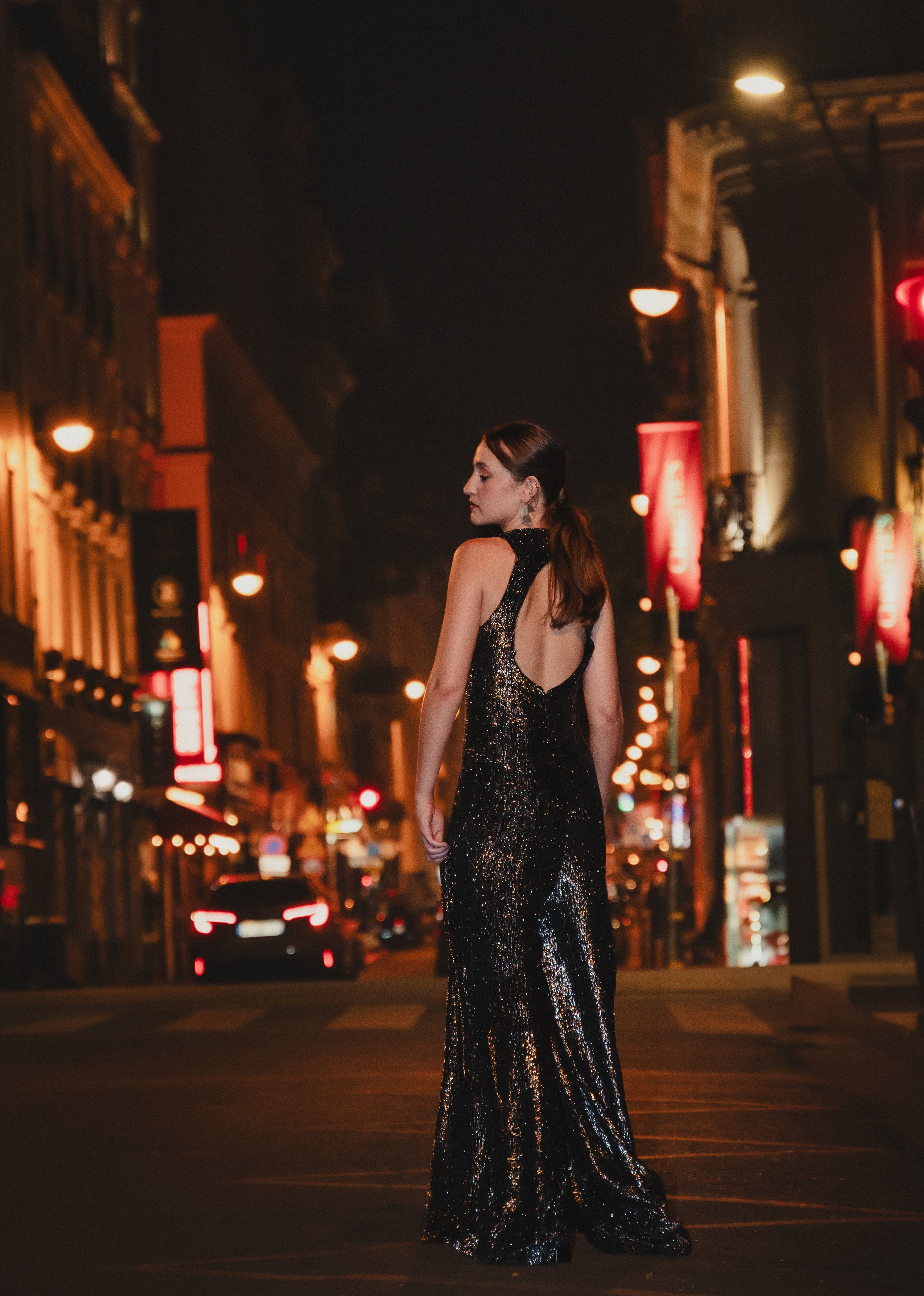 Une femme portant une robe de soirée noire brillante, debout seule dans une rue illuminée la nuit, avec des bâtiments et des enseignes lumineuses en arrière-plan.
