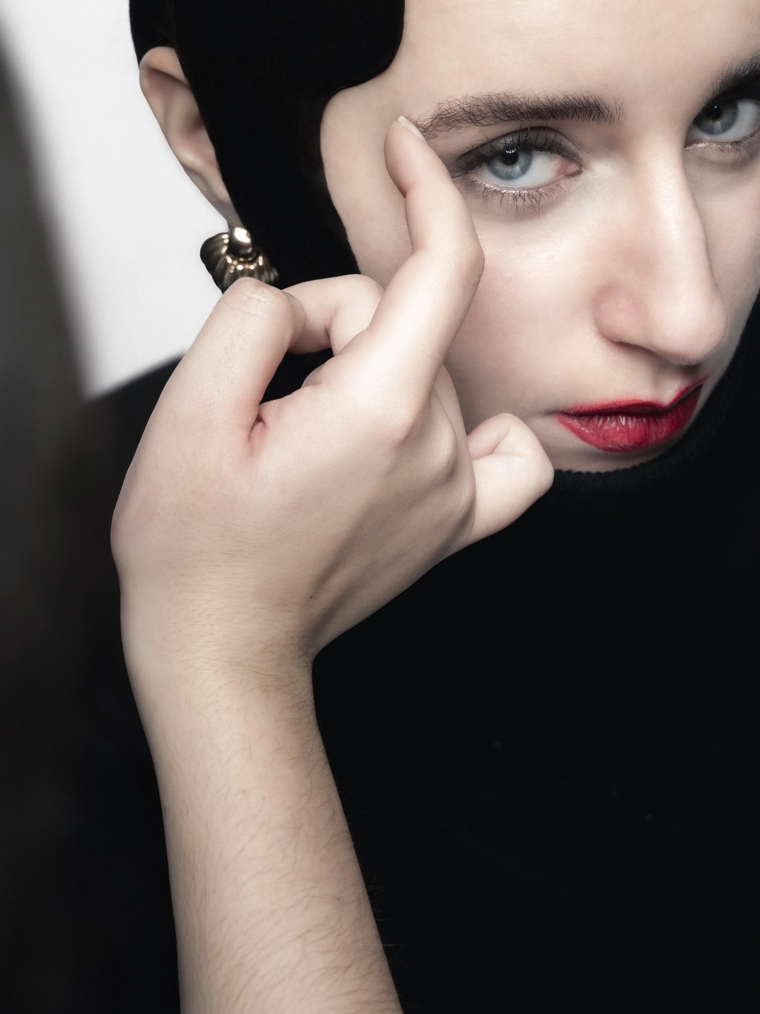 Un portrait rapproché d'une femme avec maquillage rouge, portant une boucle d'oreille en forme de coquillage, touchant son visage avec son index.