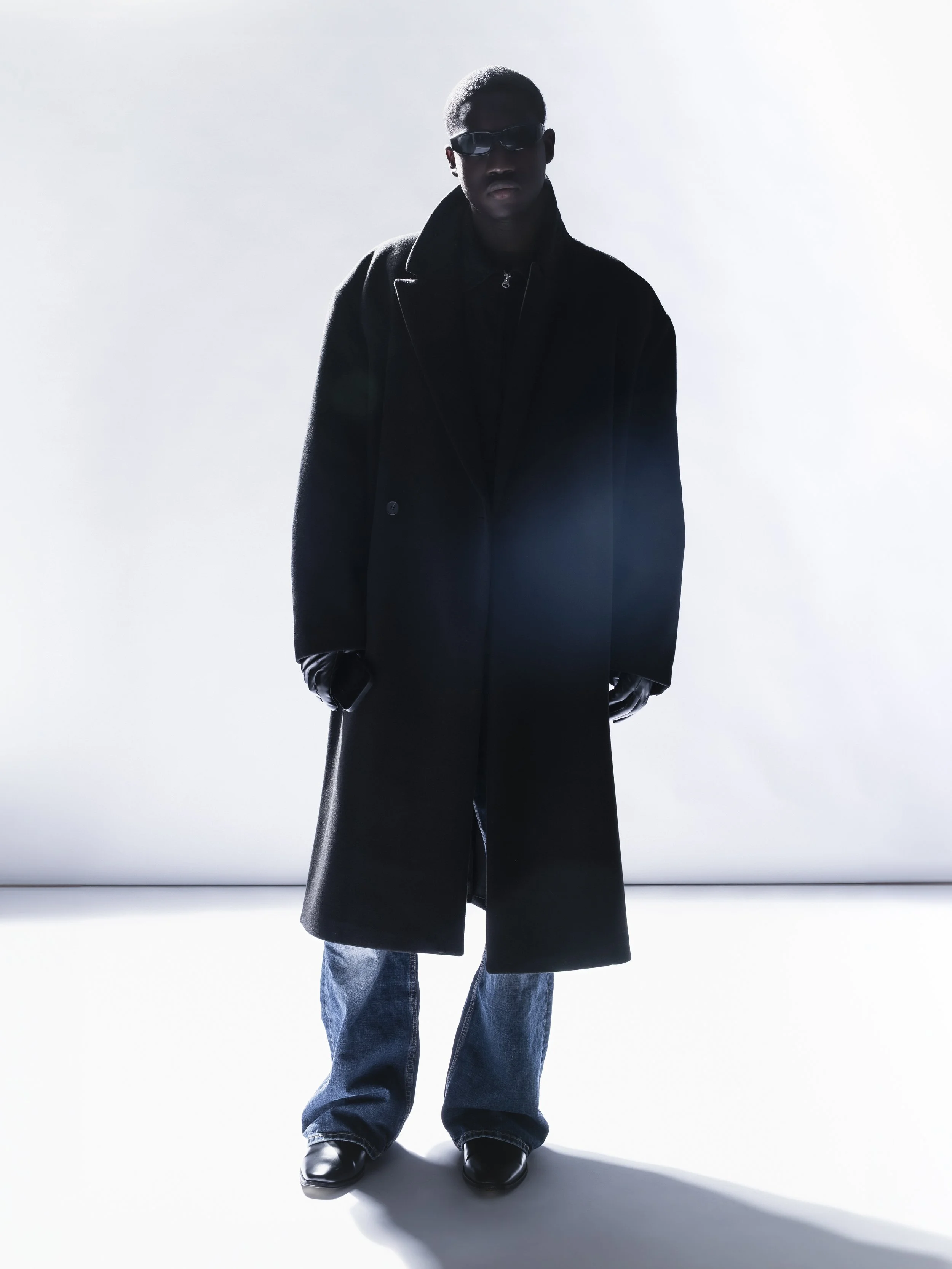 Un homme portant un manteau long noir, des lunettes de soleil, des pantalons décontractés et des chaussures noires, debout dans un fond lumineux.