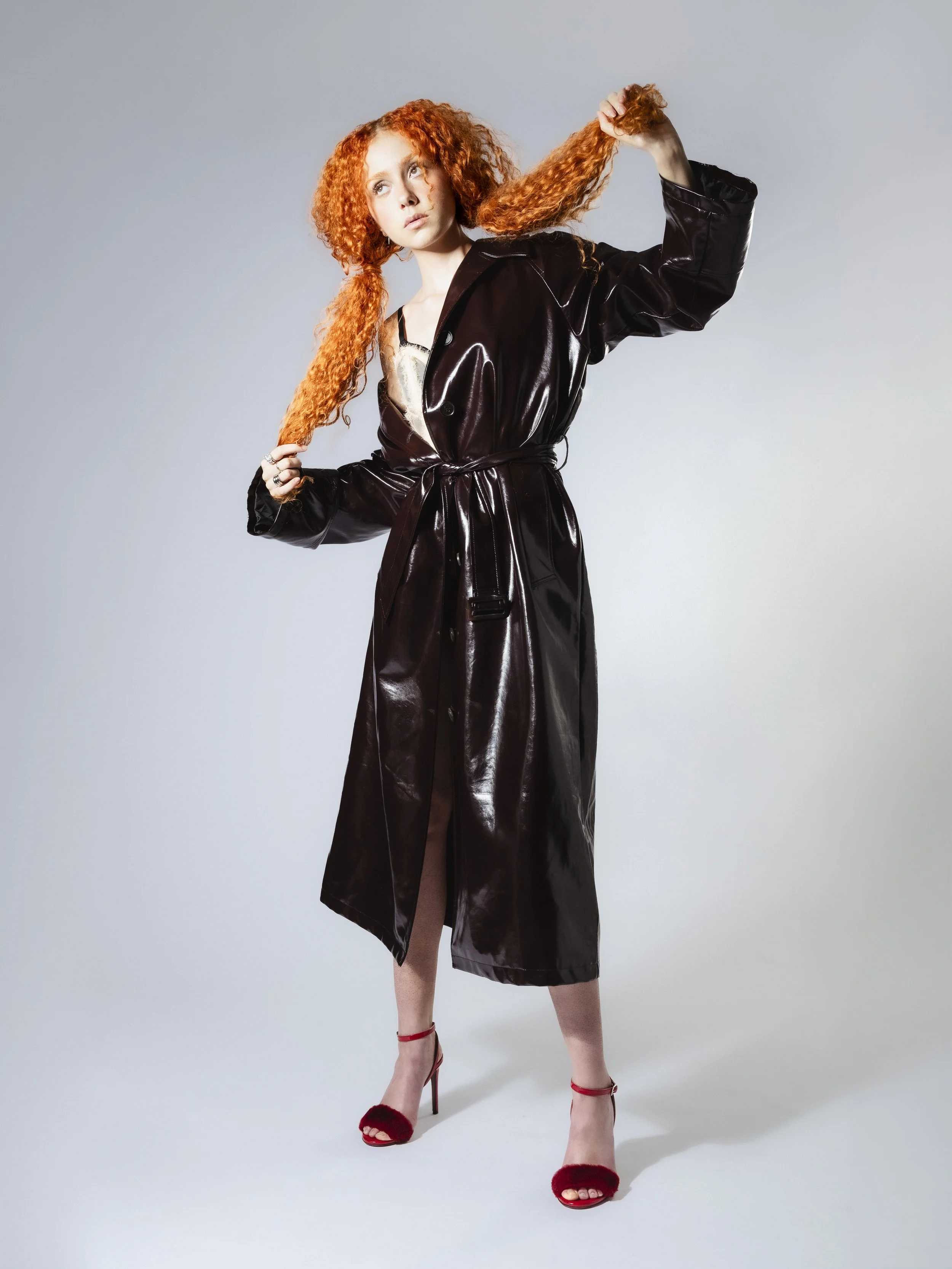 Une femme aux cheveux roux frisés portant un long manteau en vinyle noir et des talons hauts rouges pose dans un studio avec un fond gris.