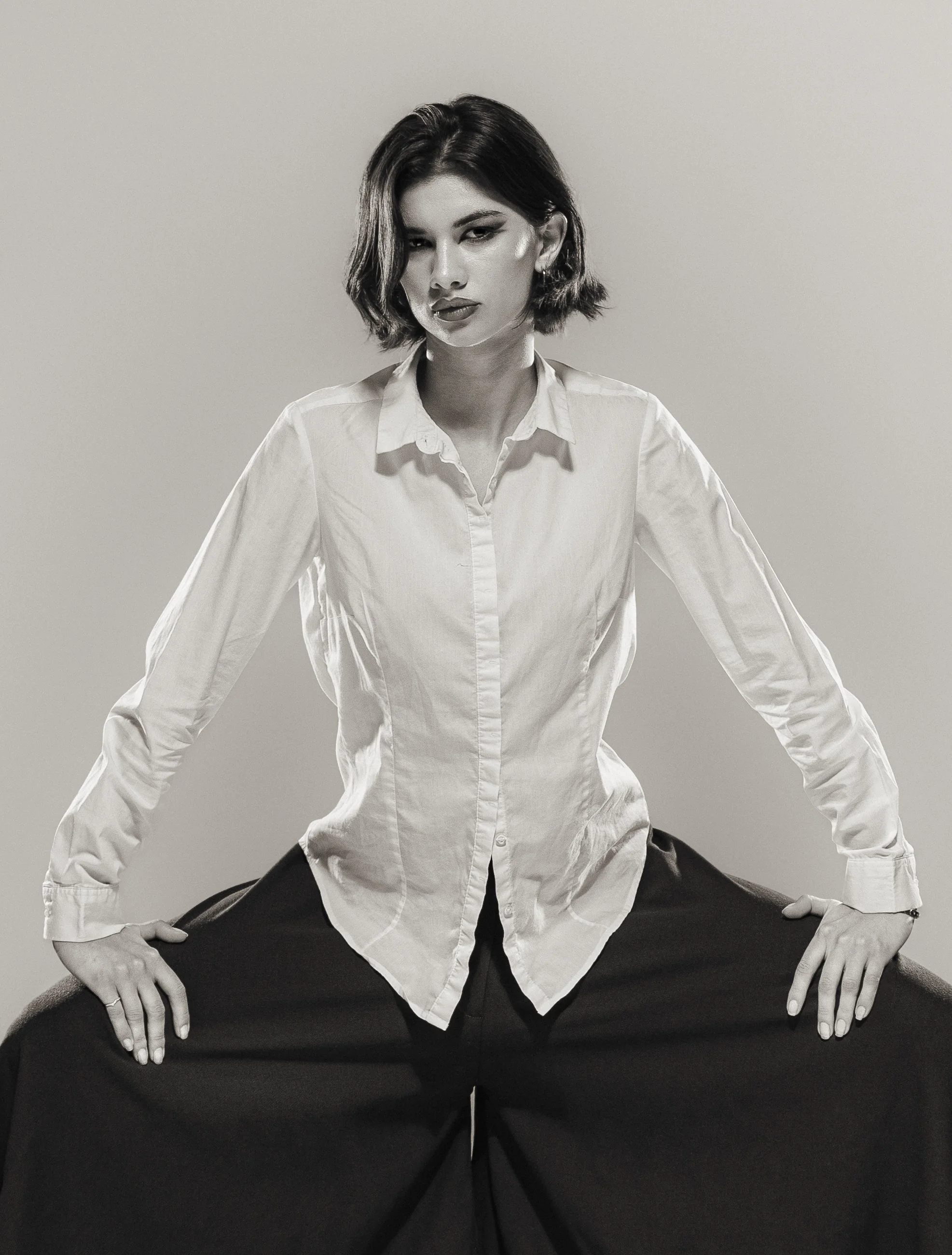 Une femme en chemise blanche et pantalon noir pose de manière confiante devant un fond neutre en noir et blanc.