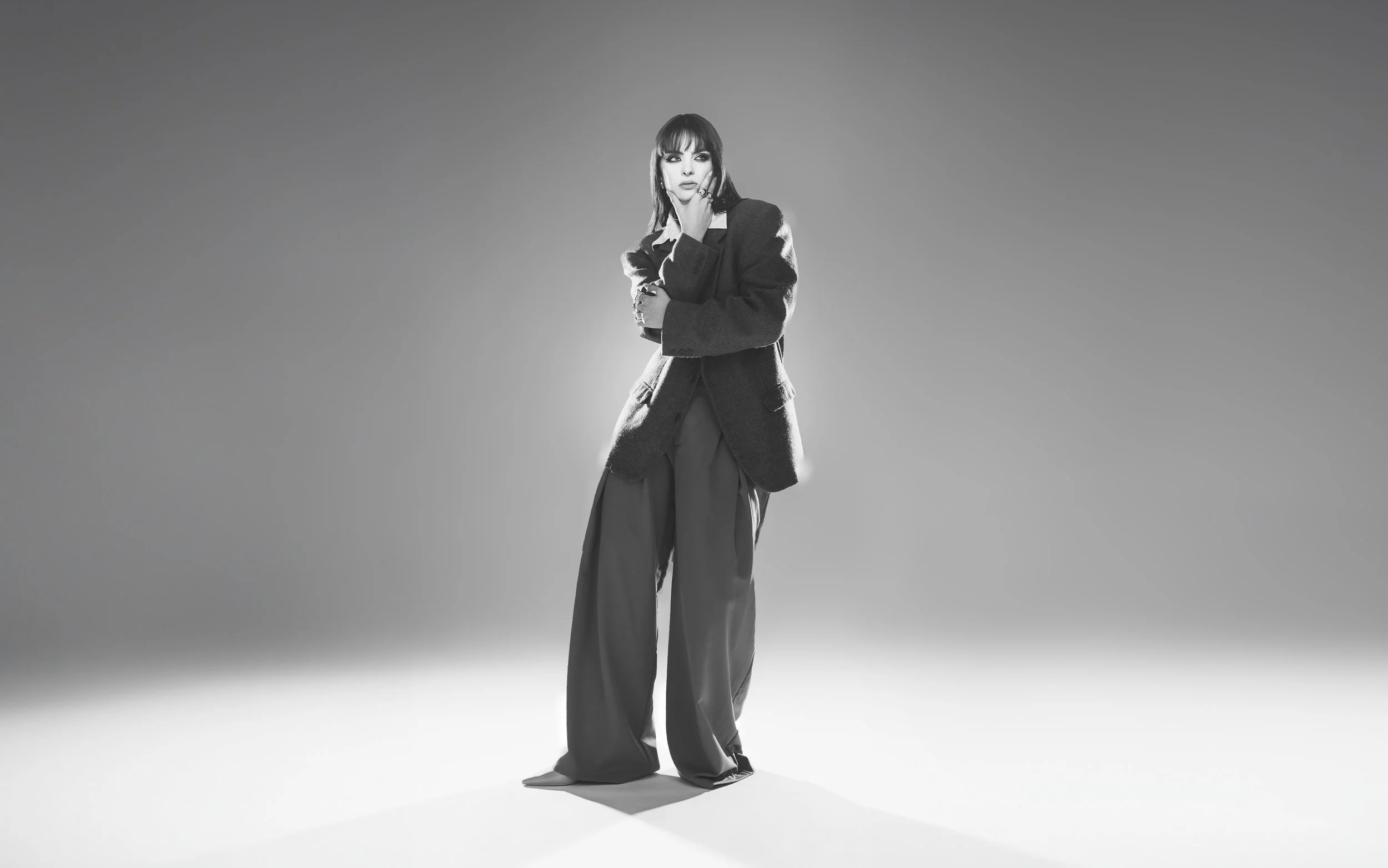Femme en vêtements amples, robe ou pantalon, avec un blazer, posant dans un studio avec un fond dégradé, en noir et blanc.