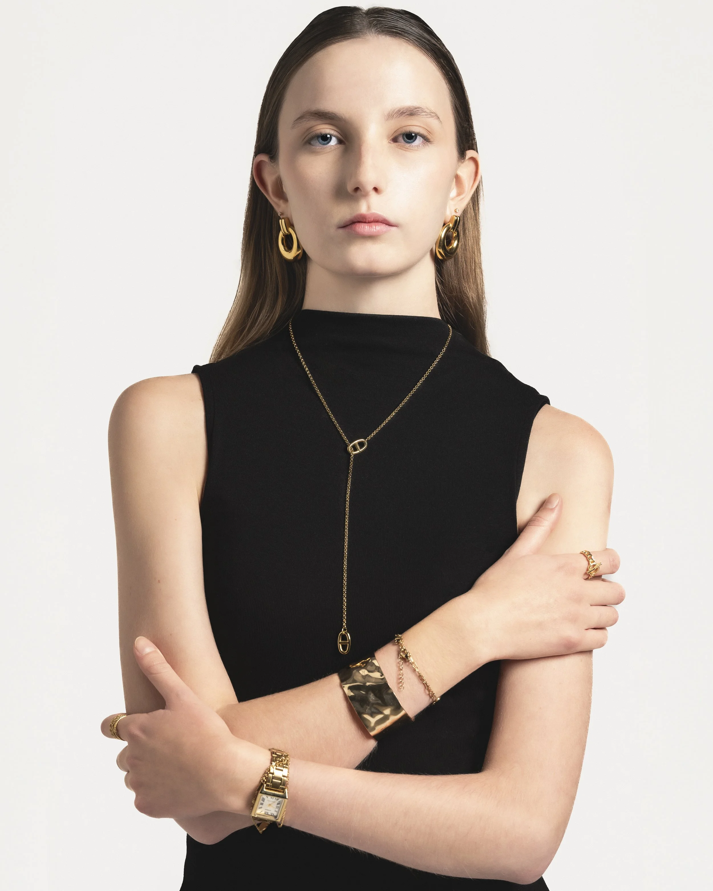 Jeune femme portant un haut noir sans manches, avec des bijoux en or, y compris des boucles d'oreilles, un collier, plusieurs bracelets, une bague, et une montre, posant les bras croisés.