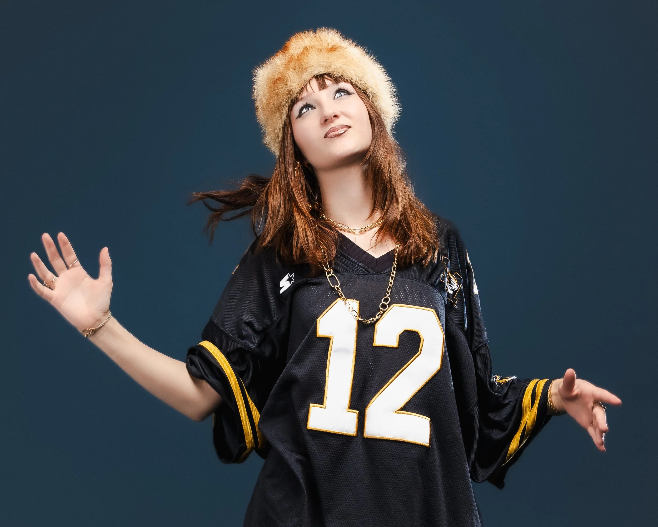 Jeune femme avec un foulard en fourrure et un maillot de football noir portant le numéro 12, posant avec des mains ouvertes contre un fond bleu.