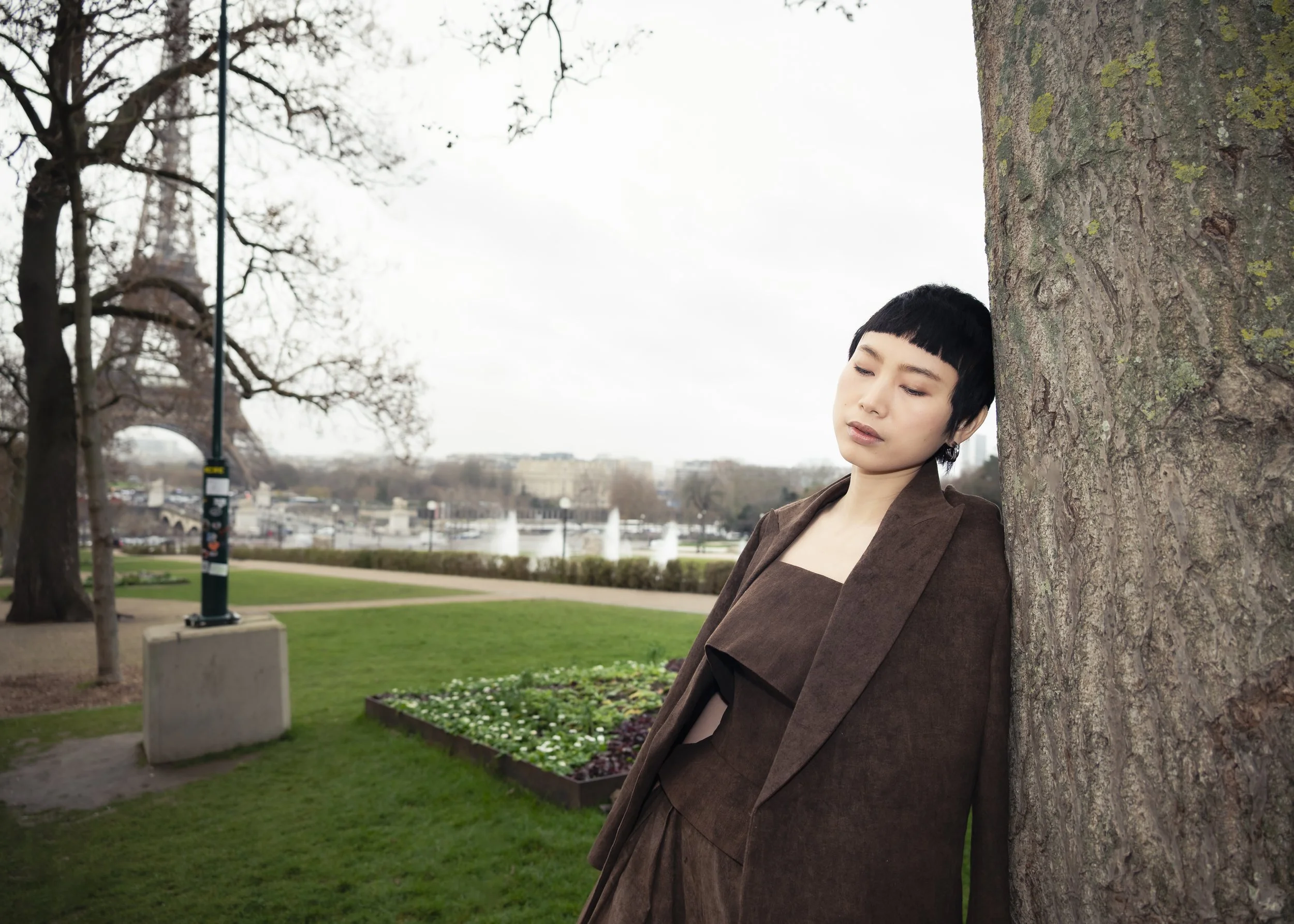 Jeune femme aux cheveux courts et noirs, portant une robe marron et un manteau, appuyée contre un arbre dans un parc, avec la Tour Eiffel et la Seine en arrière-plan, par temps nuageux.