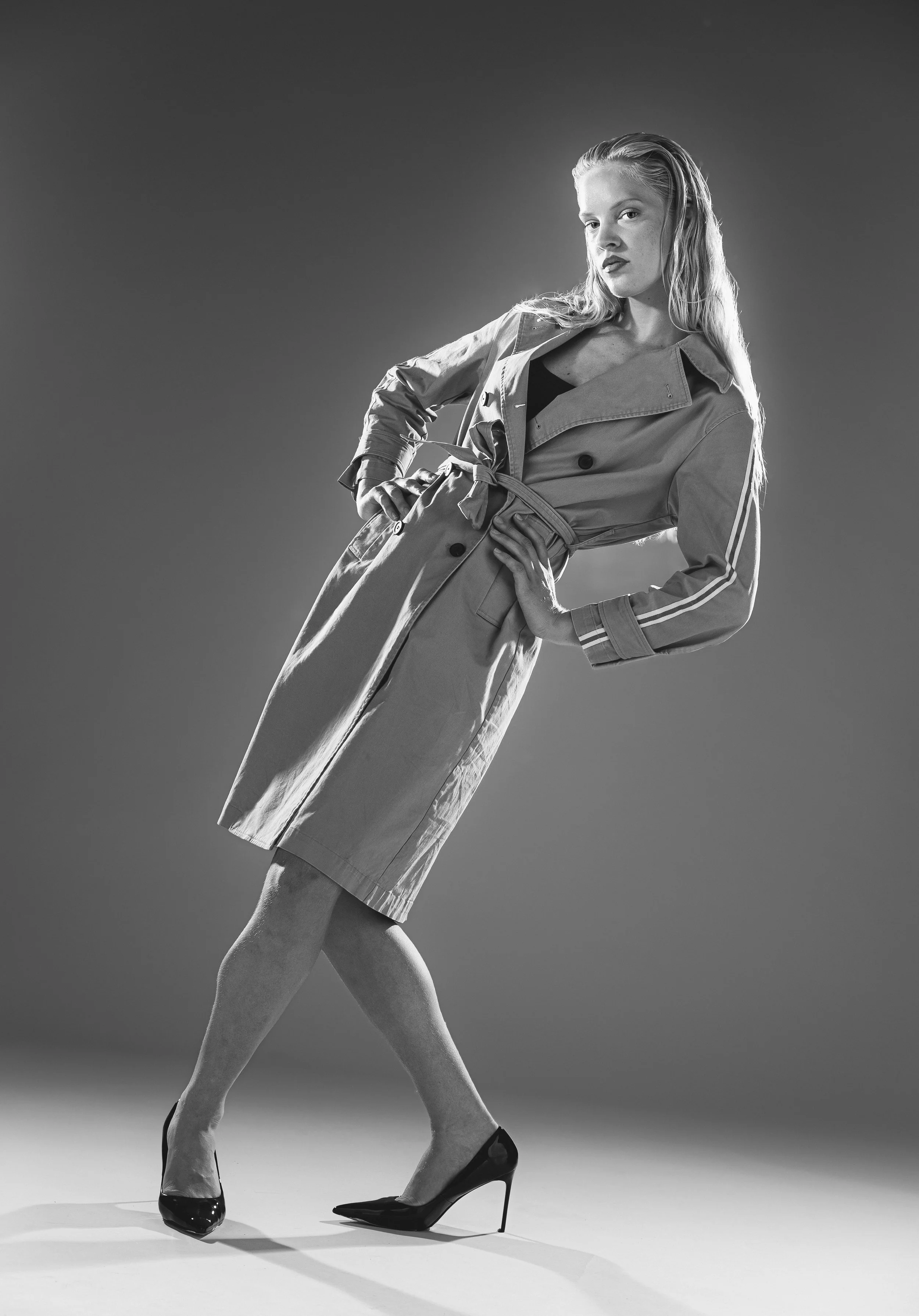 Femme en trench-coat avec haut noir, chaussure à talon haut, dans un studio, en noir et blanc.