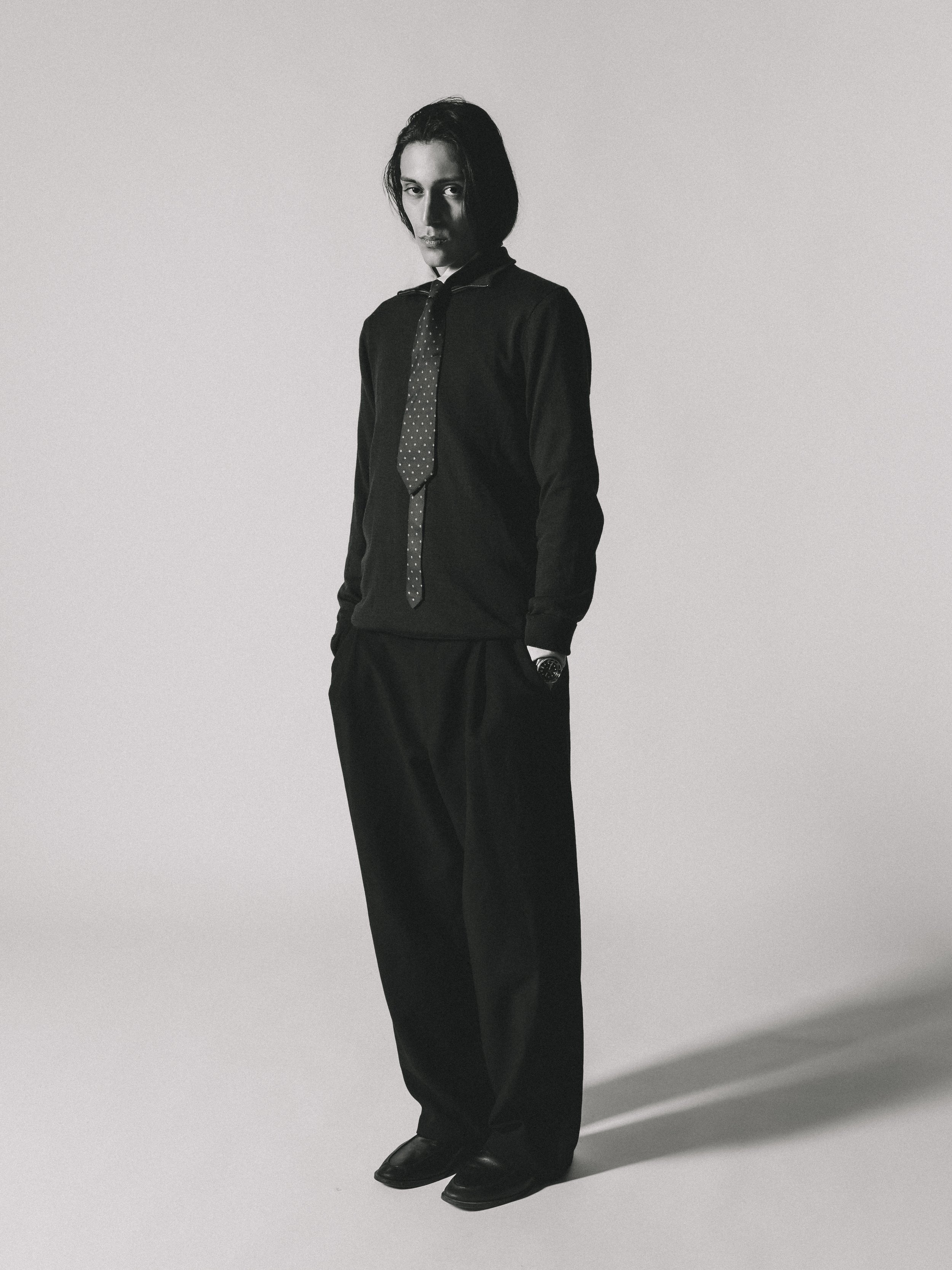 Une personne masculine habillée en noir avec une chemise, une cravate à pois, un pull et un pantalon baggy, debout contre un fond gris.