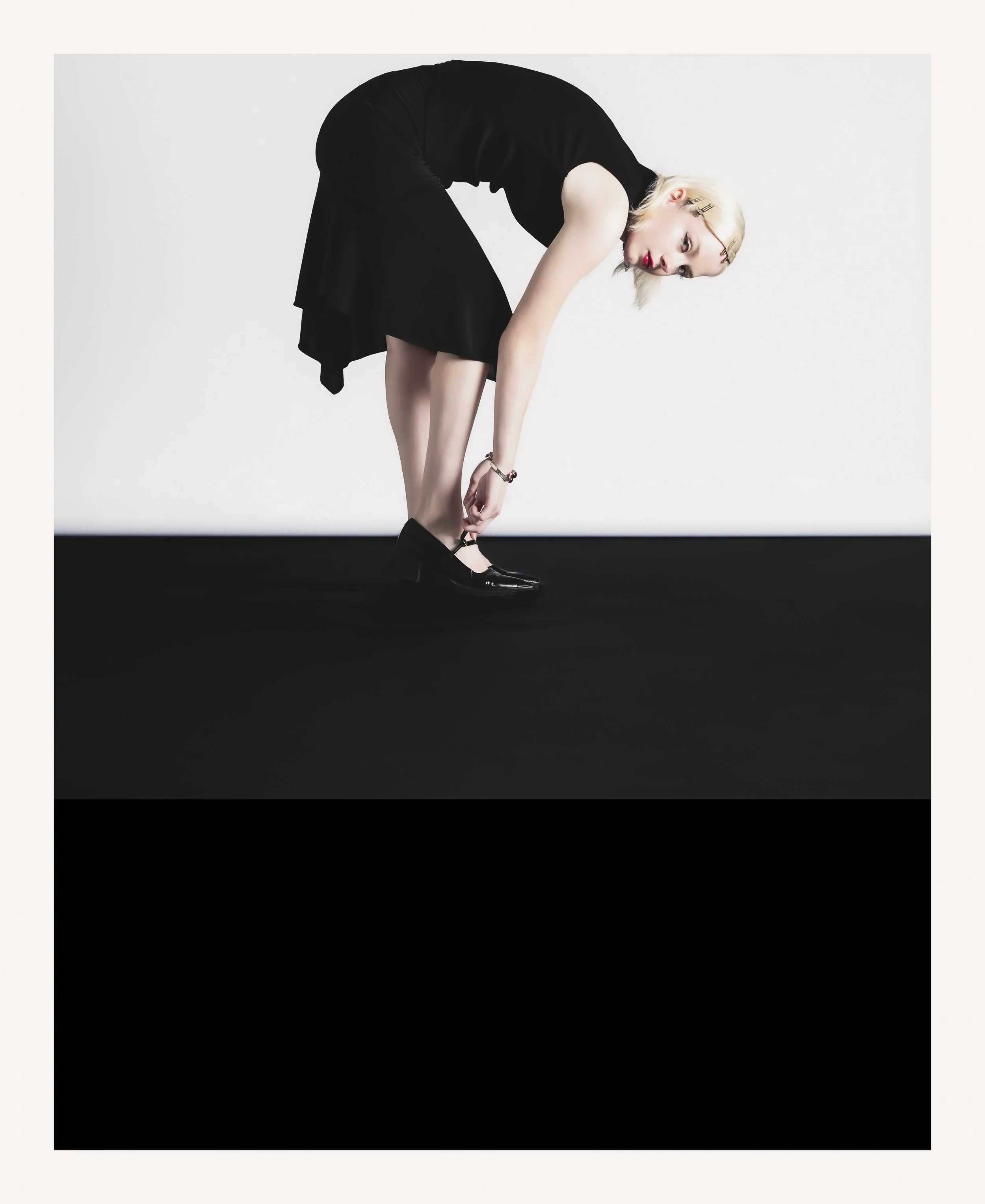 Une femme blonde habillée en noir, pliée en avant en train de boutonner ses chaussures à talons noirs, avec un fond blanc et noir.