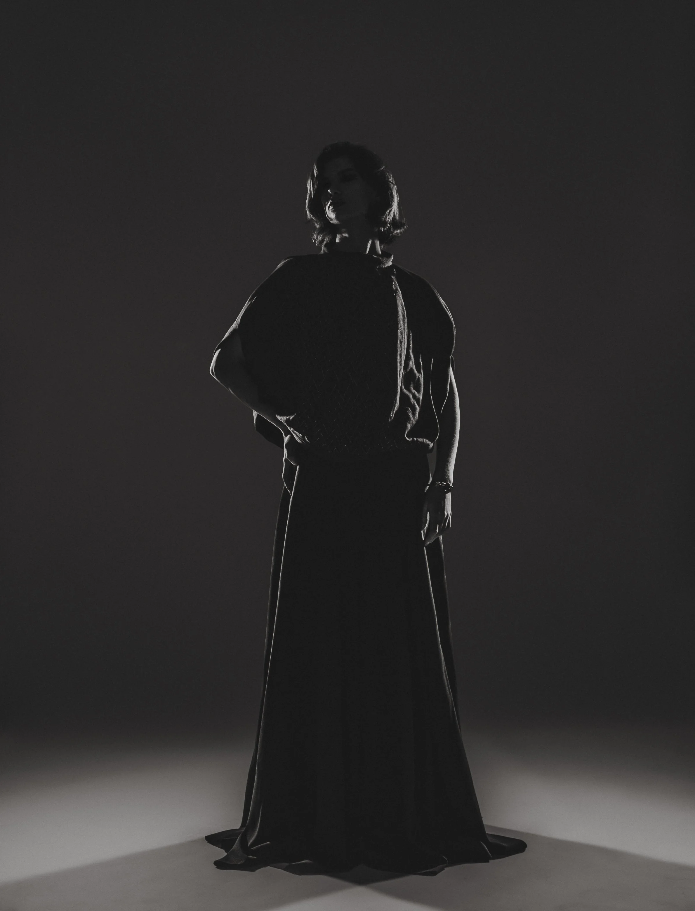 Femme en silhouette noire et blanc, debout, vêtue d'une robe longue, dans un éclairage dramatique à contraste élevé.