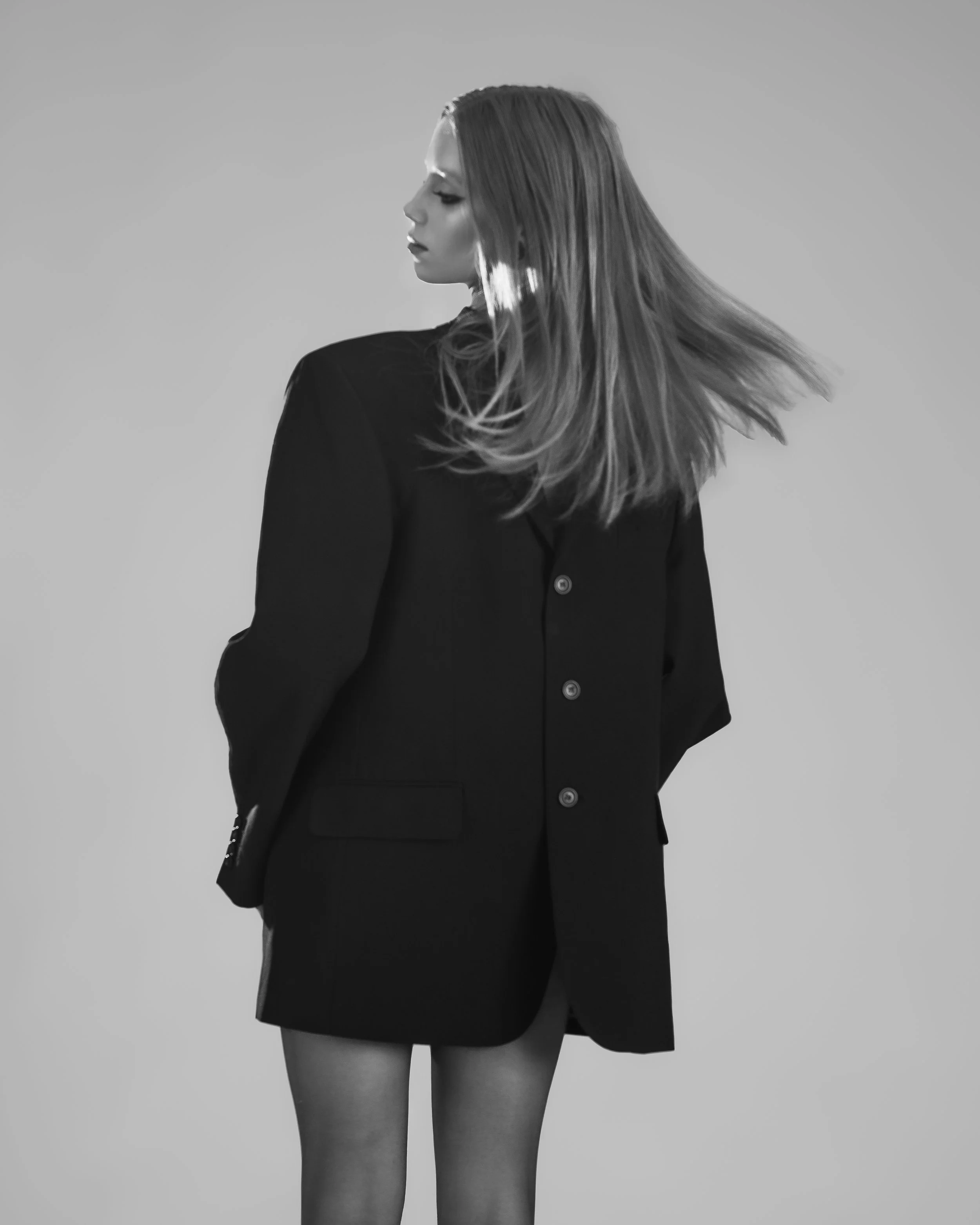 Femme portant un tailleur noir, vue de dos, cheveux longs et raides, fond gris.