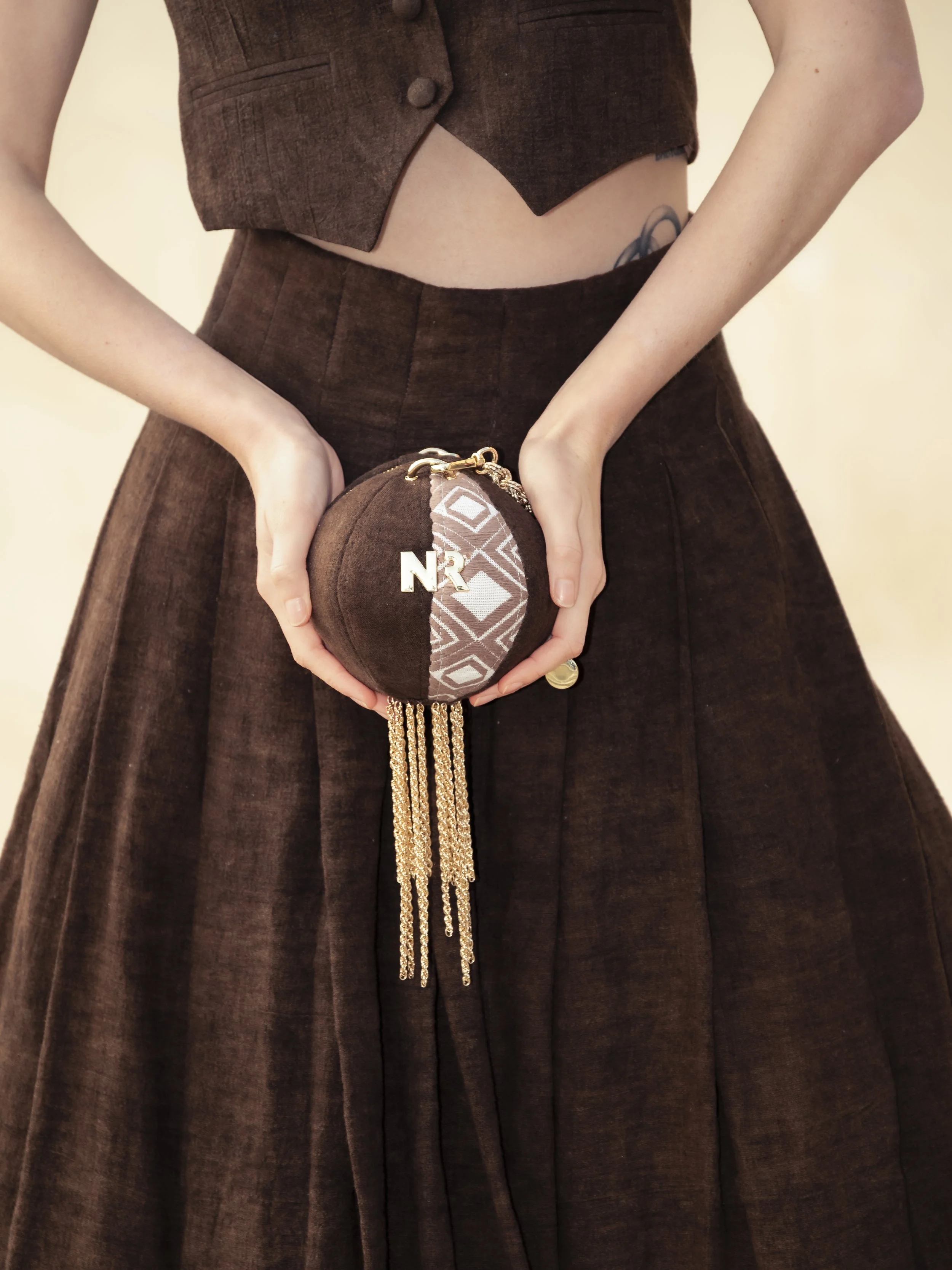 Une femme porte une robe marron foncé et tient un sac à main rond, décoré de chaînes dorées, avec des lettres N et R en métal, et un motif géométrique. La photo montre le haut de la robe et le sac.