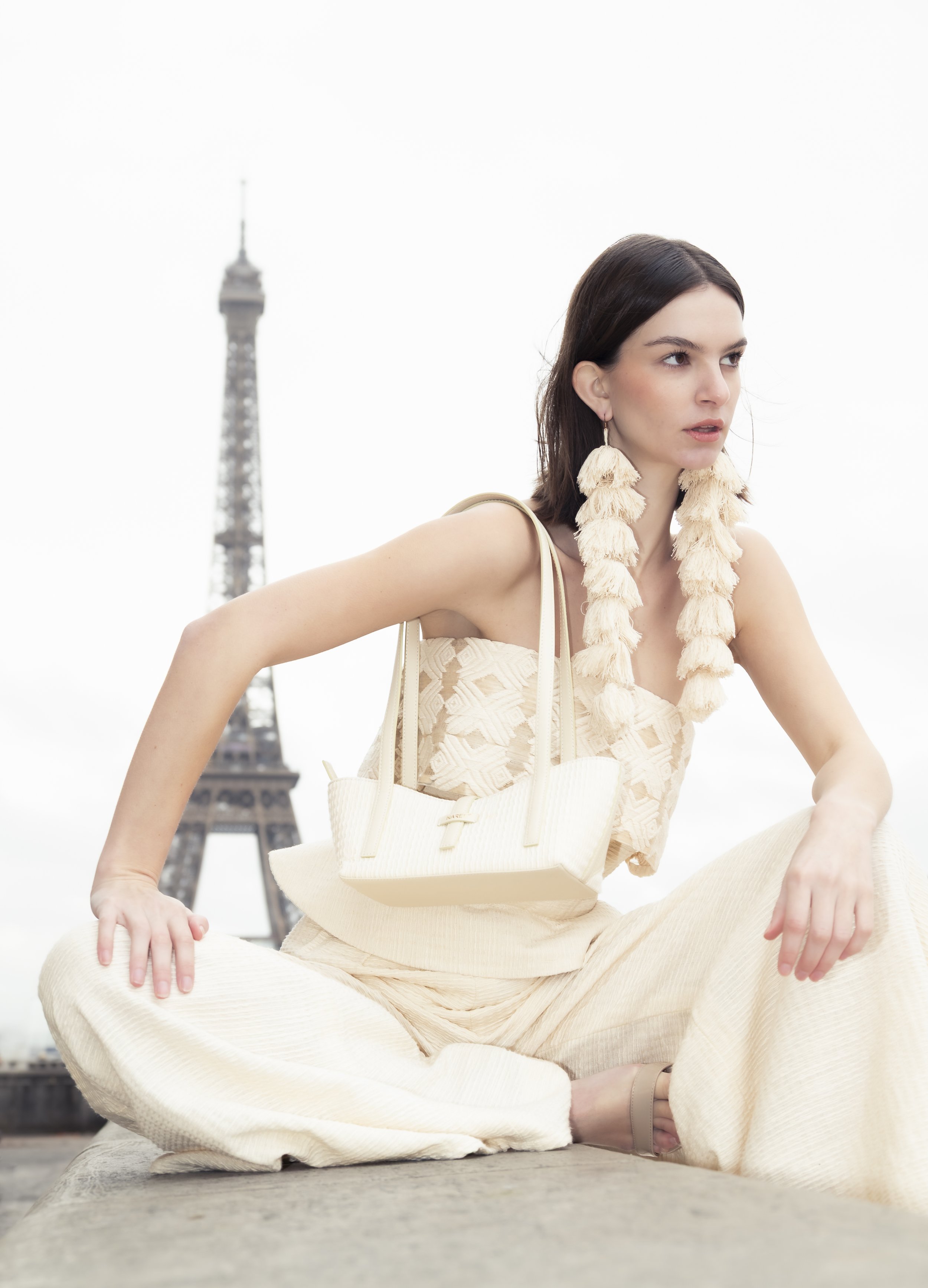 Jeune femme en tenue beige assise sur une surface avec la Tour Eiffel en arrière-plan, portant de longues boucles d'oreilles en plumes et un sac beige.